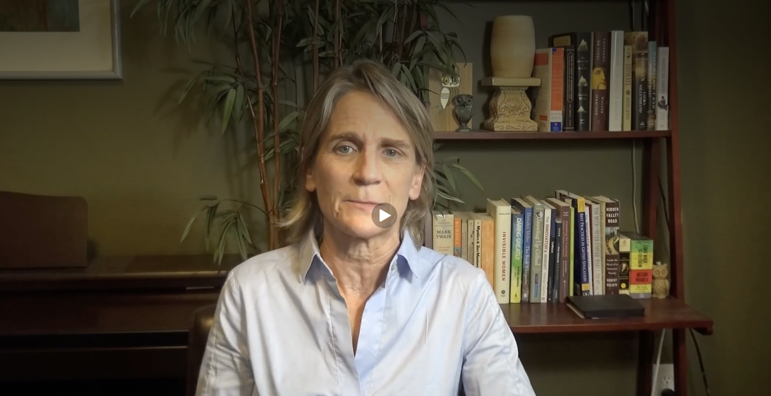 A middle-aged woman with shoulder-length blonde hair, wearing a light blue collared shirt, sitting in front of a bookshelf filled with various books and decorative items, in a room with green walls and houseplants.
