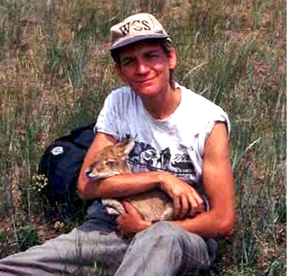 Sharon Deem wearing a white cap, gray t-shirt, and carrying a backpack, sitting outdoors on grass while holding a small dog.