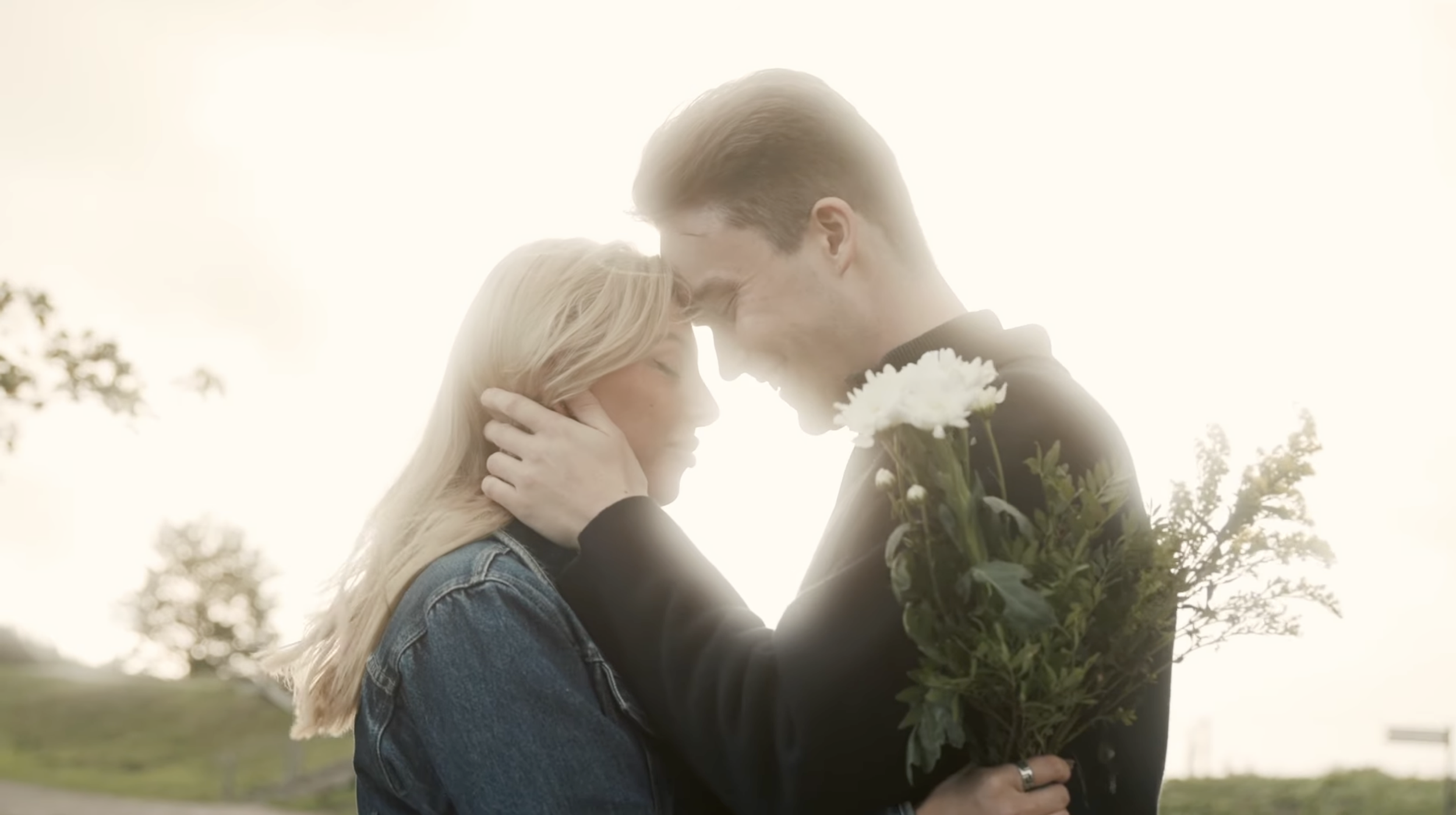 A man and a woman embrace outdoors at sunset, with the woman holding a bouquet of white flowers.