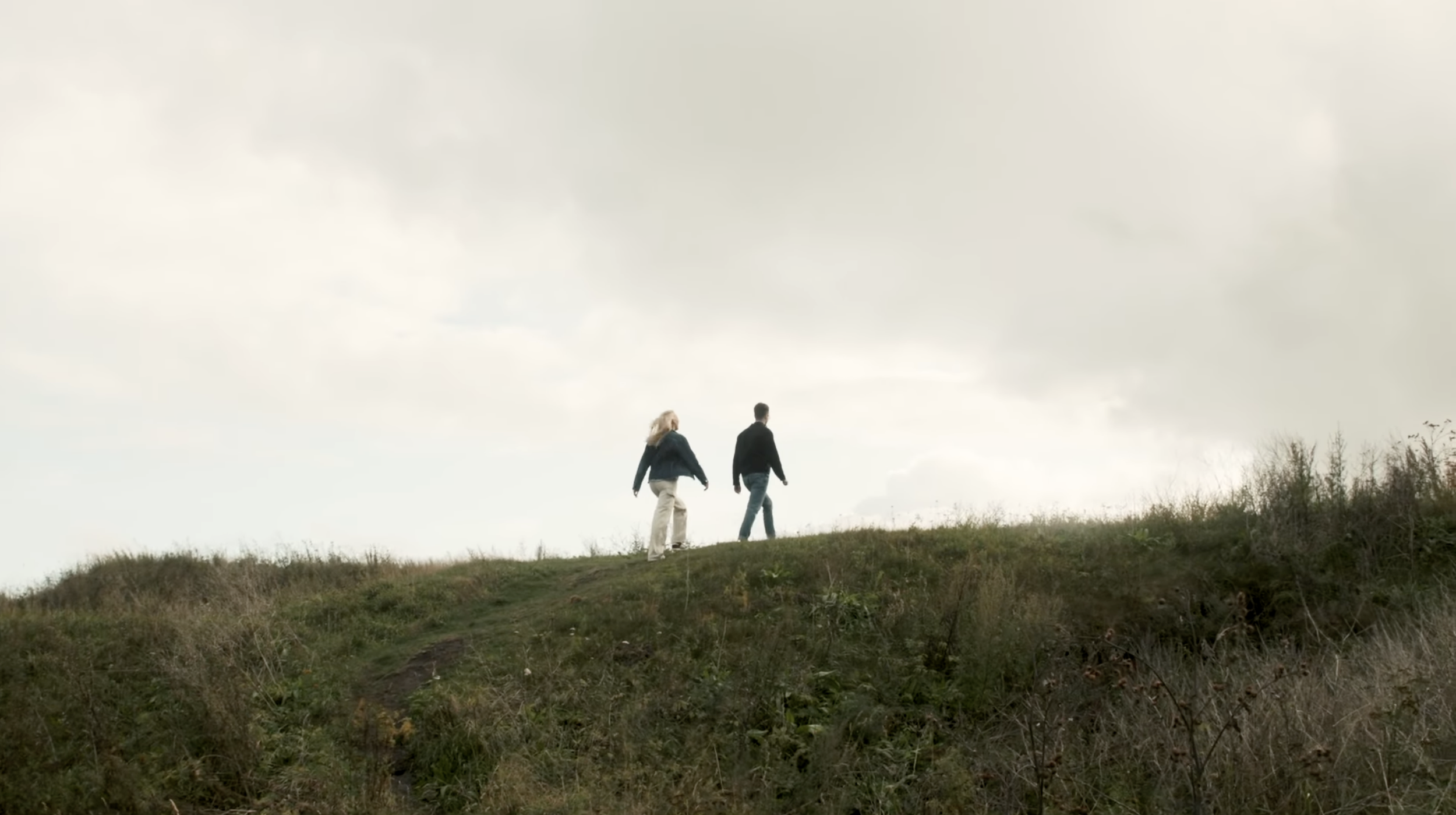 Two people, a woman and a man, walking up a grassy hill under a cloudy sky.