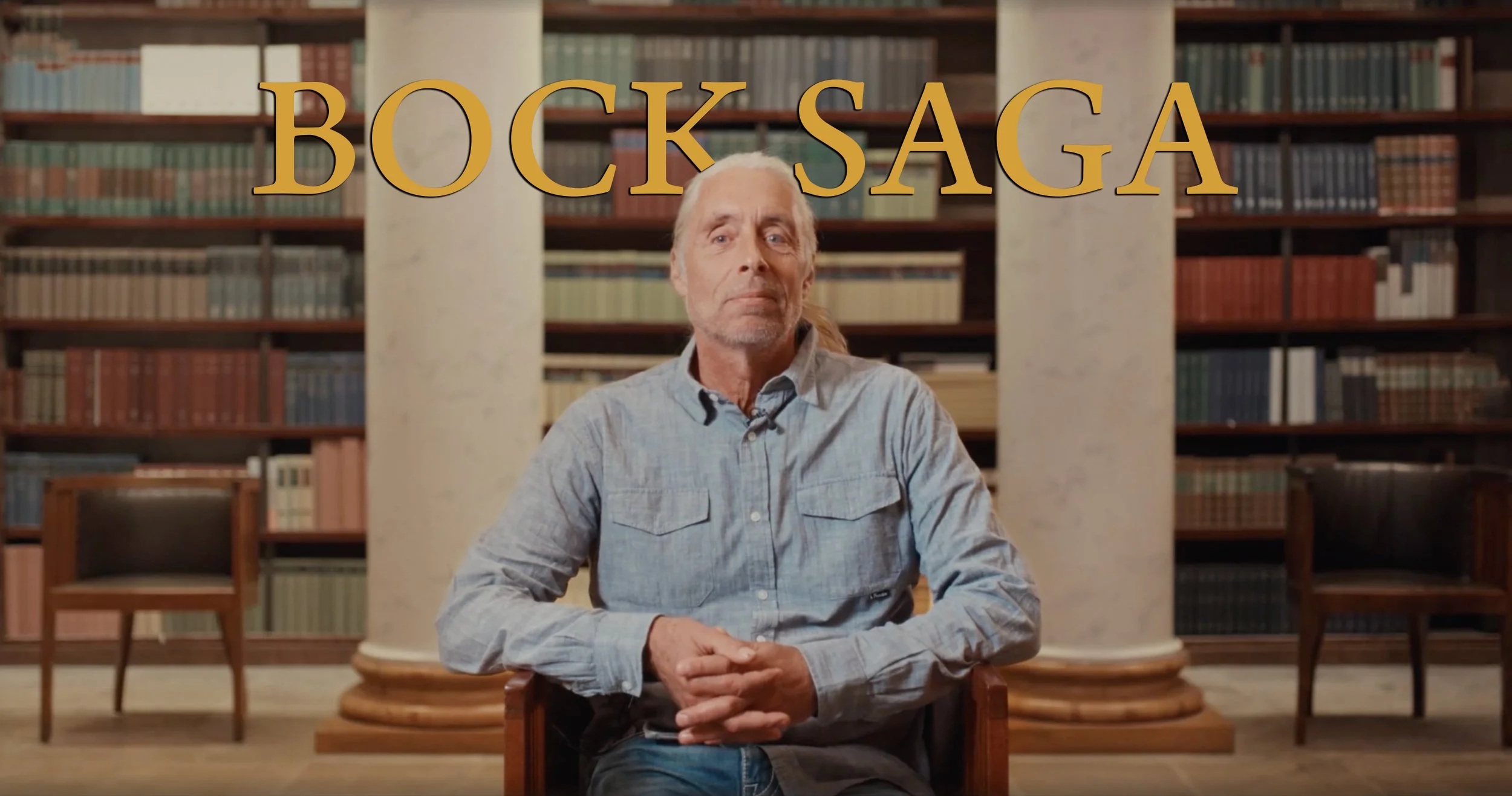 A man with long hair and a beard sitting in front of a bookshelf in a library or study, with the words "BOOK SAGA" overlaid in large yellow text.