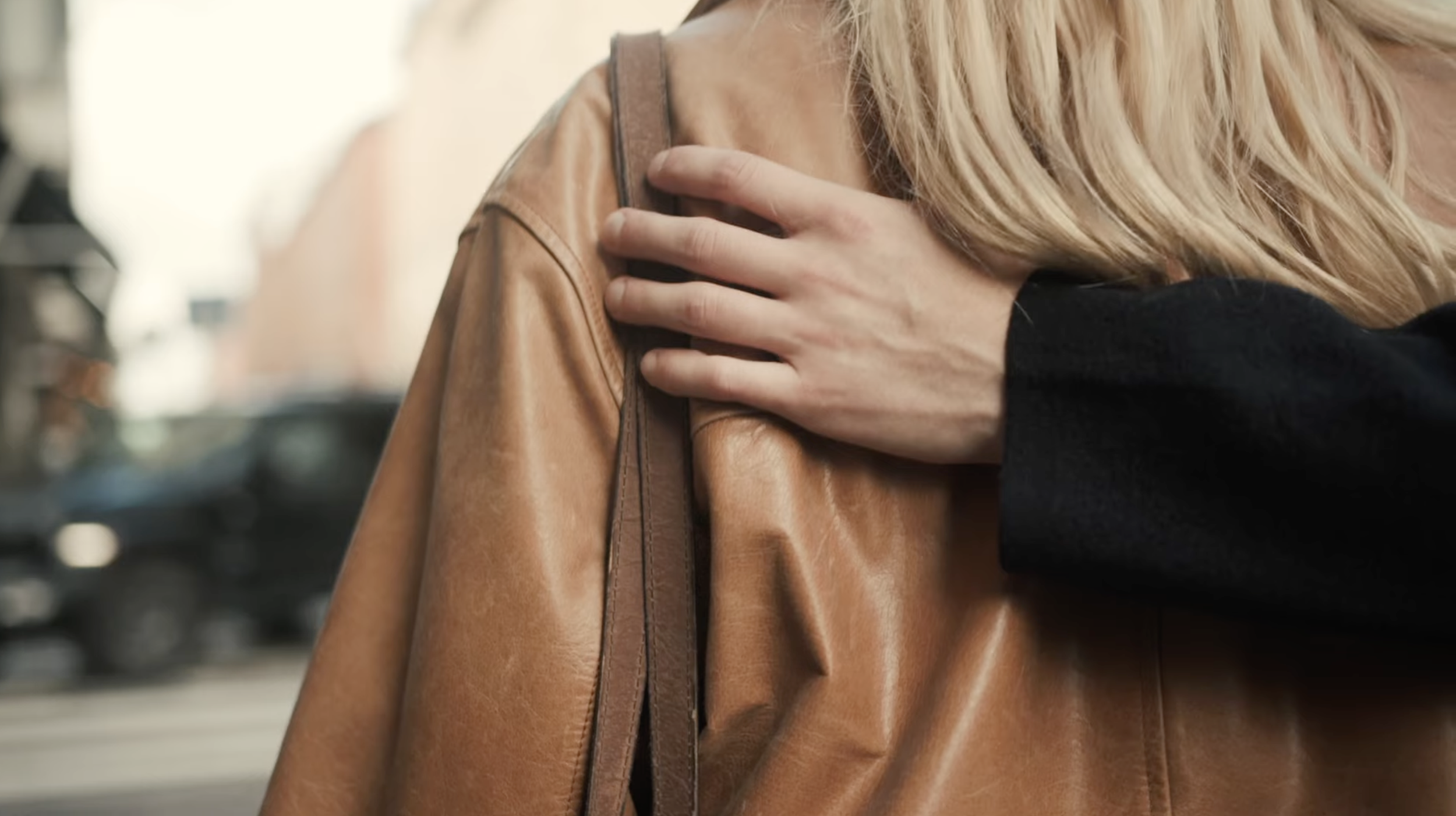 A person with blonde hair wearing a black top, holding a tan leather bag on their shoulder, with an urban street background.