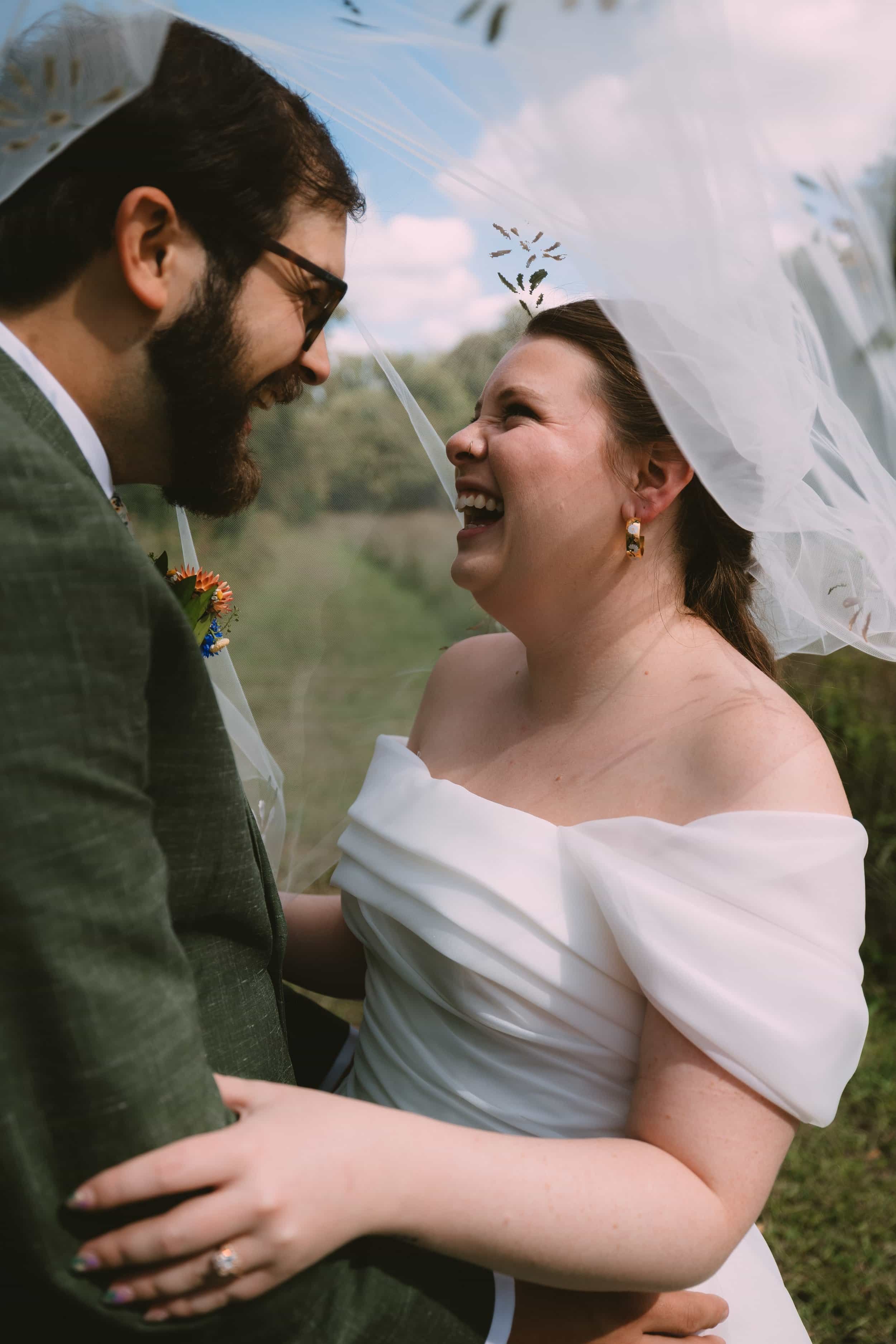 A bride and groom smiling and holding each other under a sheer veil outdoors during their wedding.