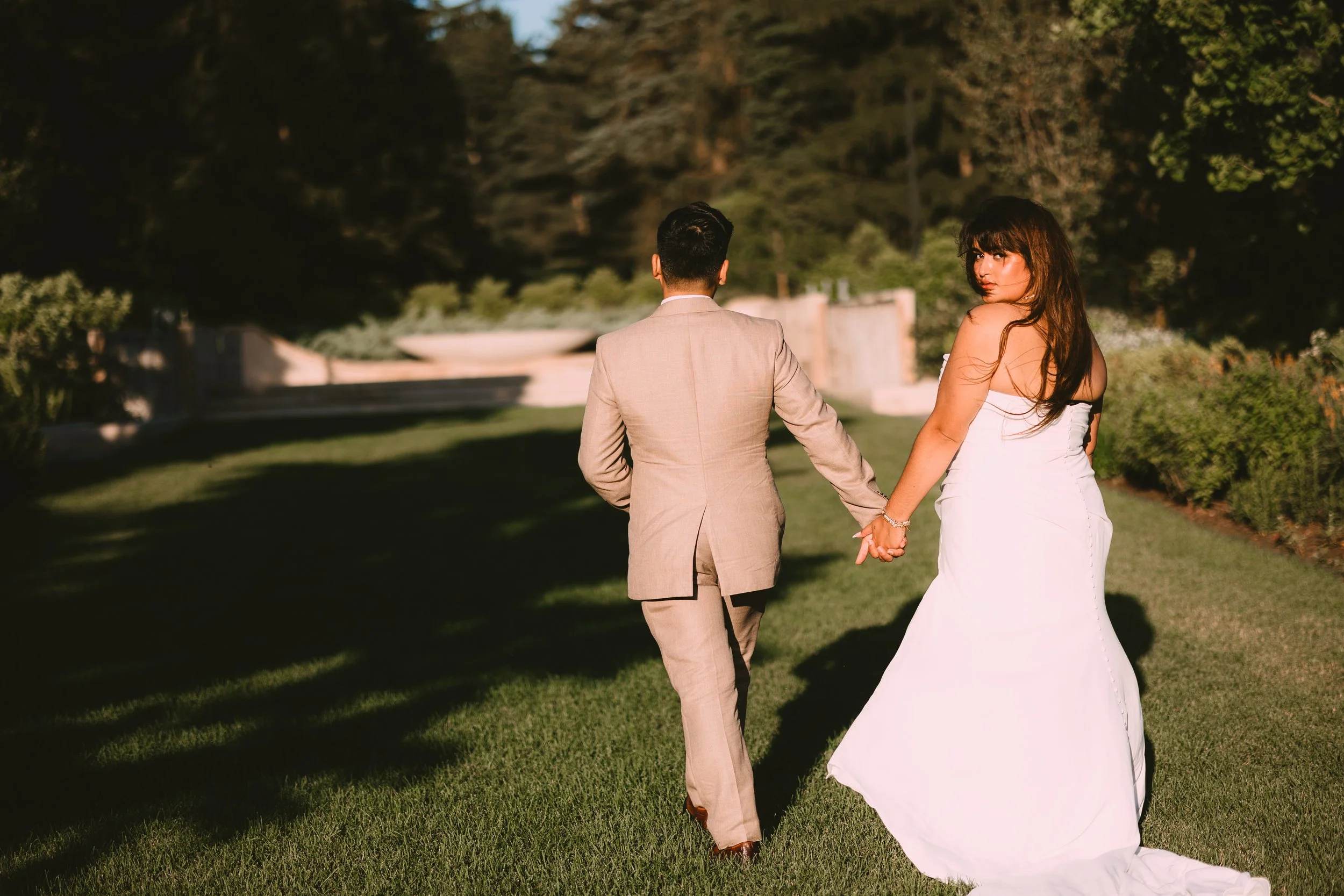 A man and woman in formal attire walking hand in hand on a grassy lawn during sunset, with trees and a fountain in the background.