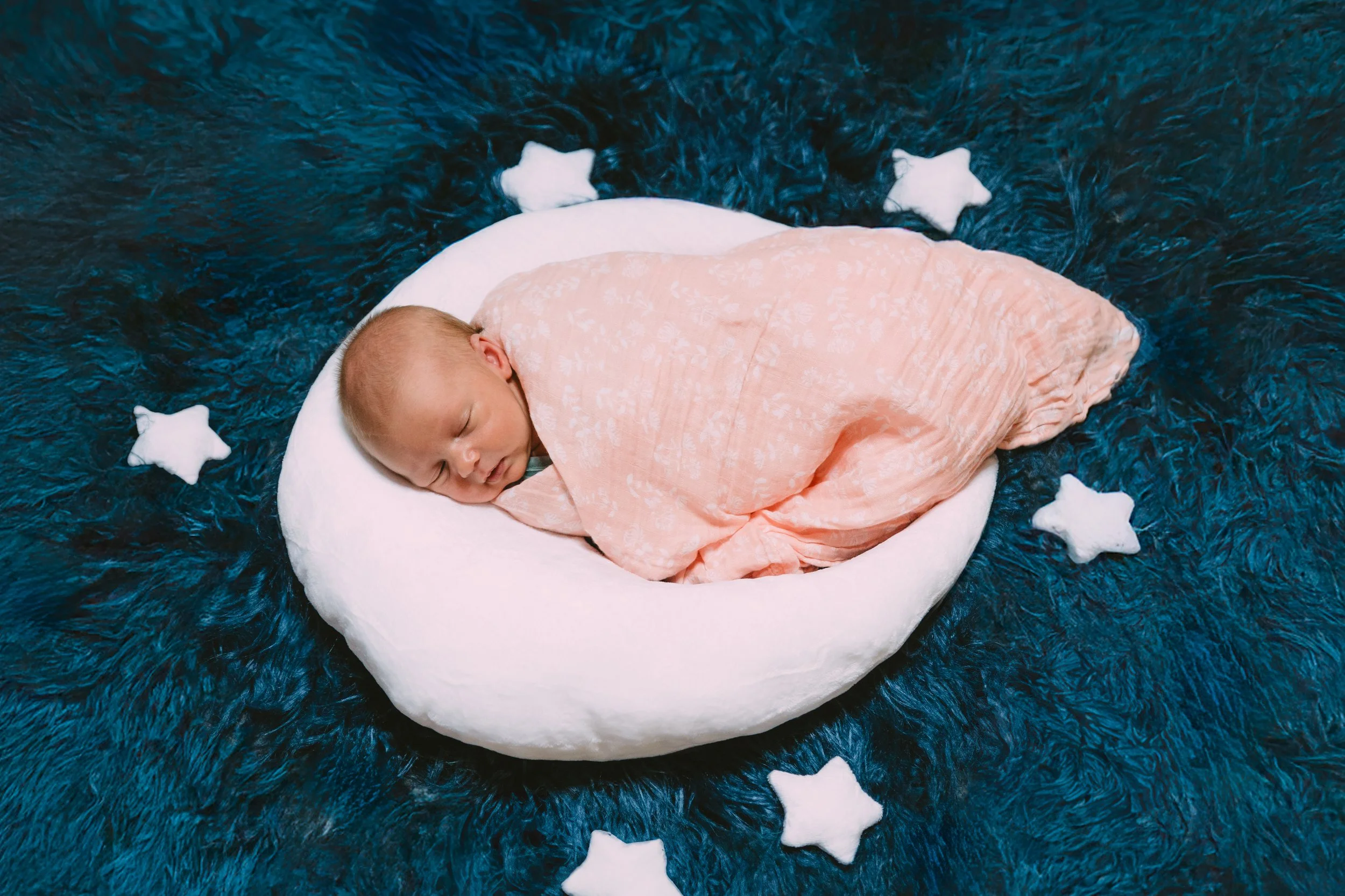 A sleeping baby wrapped in a peach blanket, lying on a white circular cushion on a dark blue furry blanket, with small white star-shaped pillows arranged around the cushion.