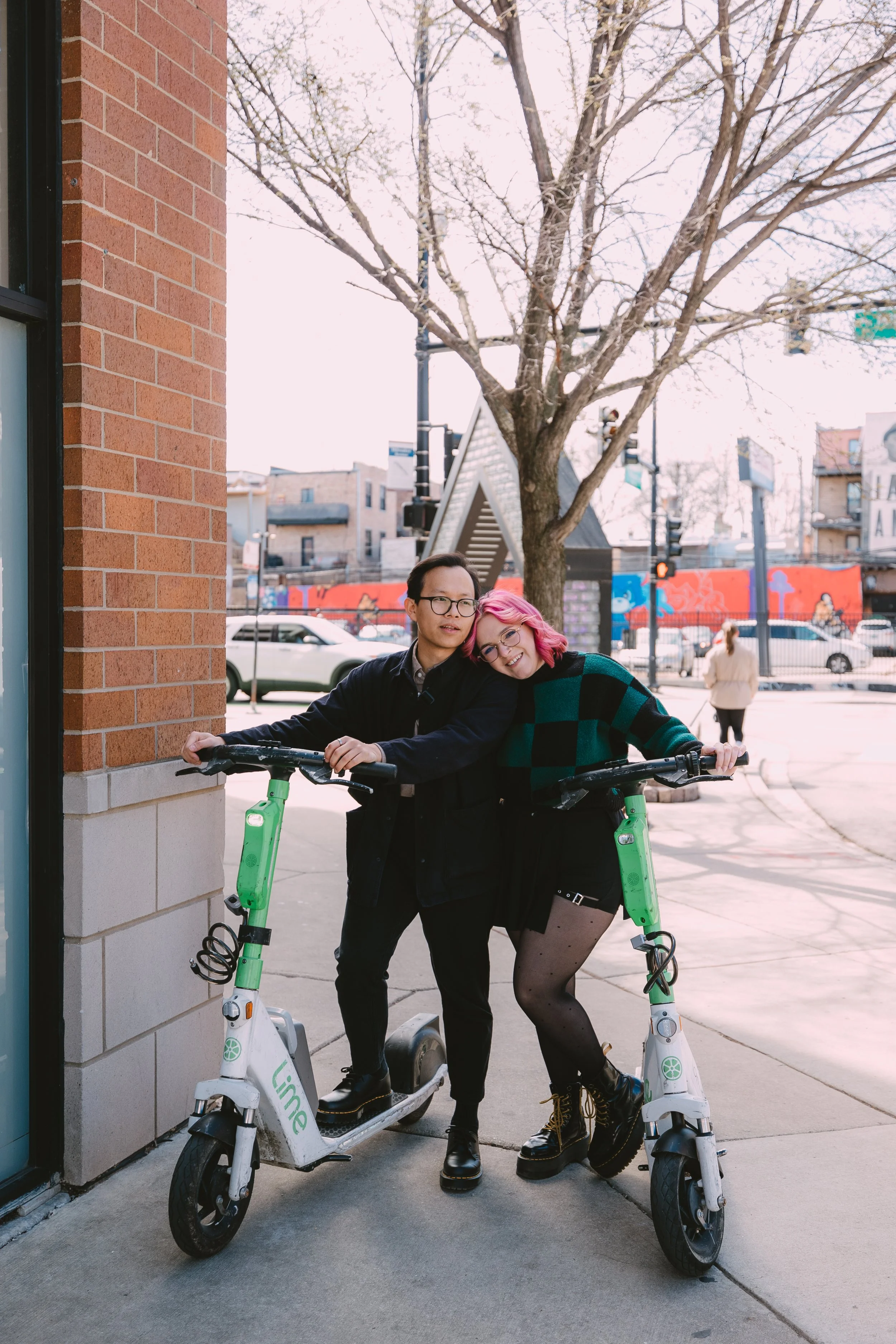 A young man and woman stand outside next to two Lime electric scooters, embracing and smiling. The woman leans her head on the man's shoulder. The background features a leafless tree, cars, and a colorful mural on buildings.