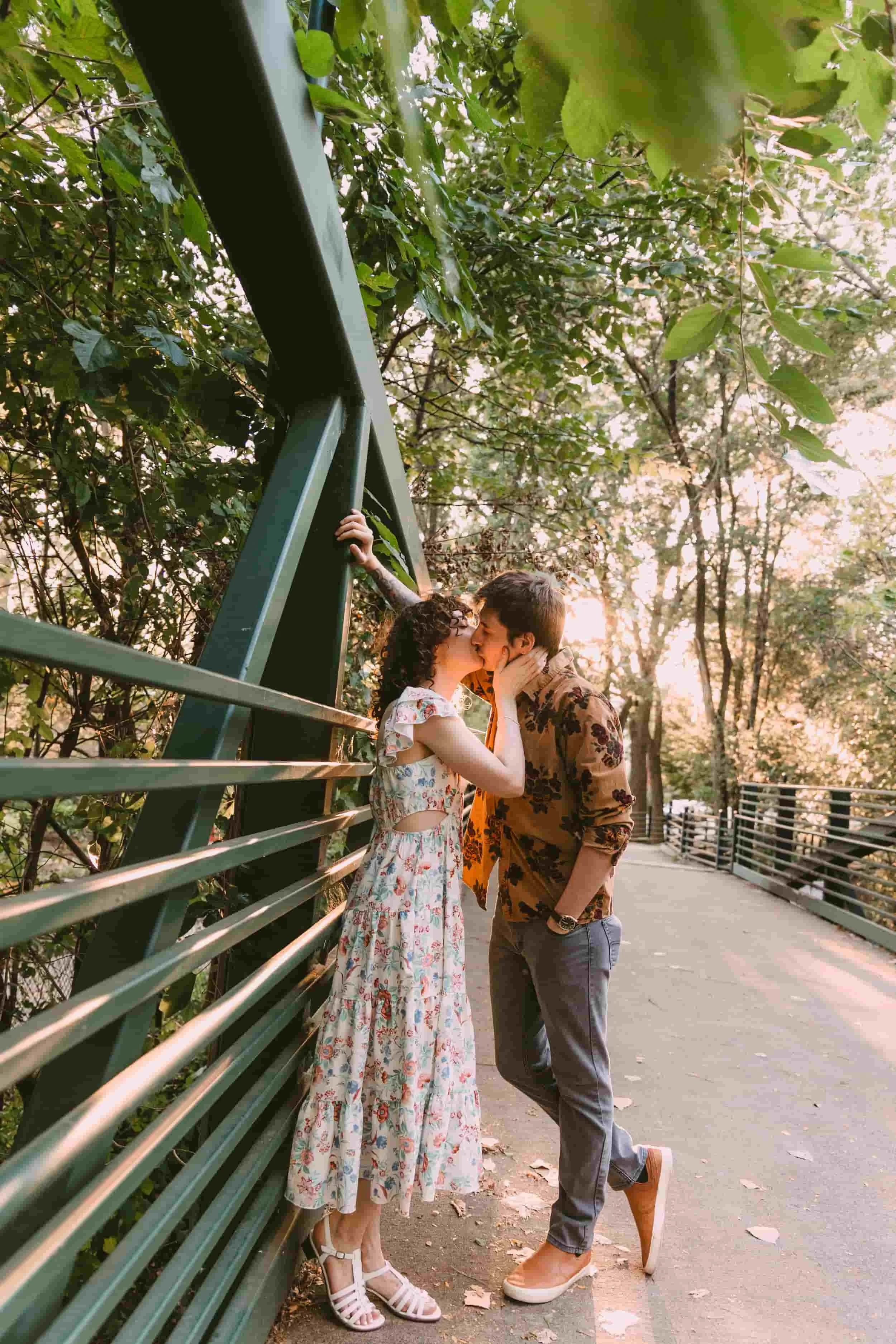 A couple sharing a kiss on a park pathway during sunset, with a green metal fence and lush trees in the background.