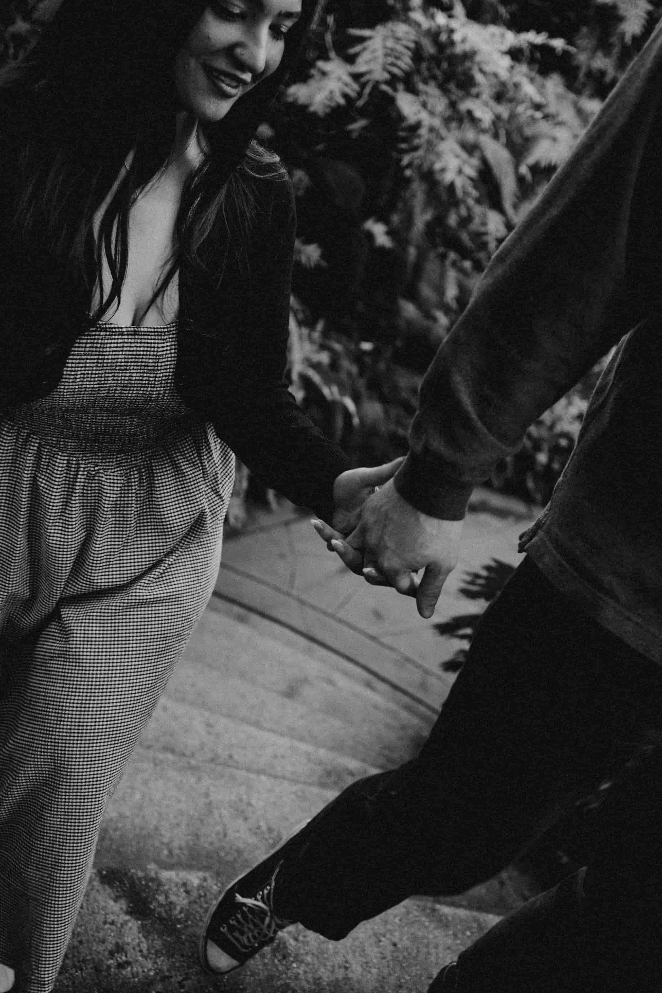 A woman and a man holding hands, standing outdoors on a sidewalk, with foliage in the background.