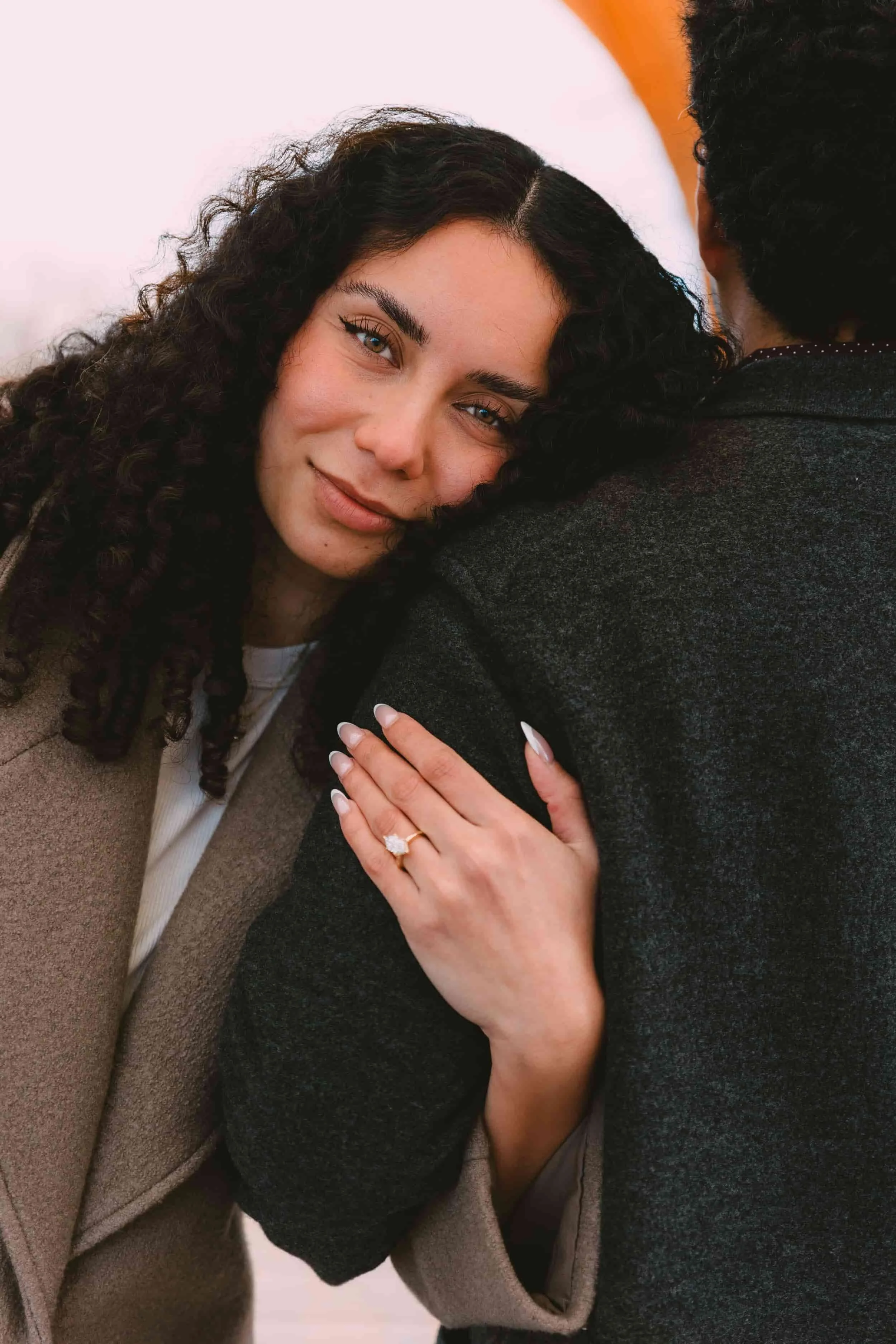 A woman with dark curly hair, smiling gently, resting her head on a man's shoulder, showing an engagement ring on her left hand.