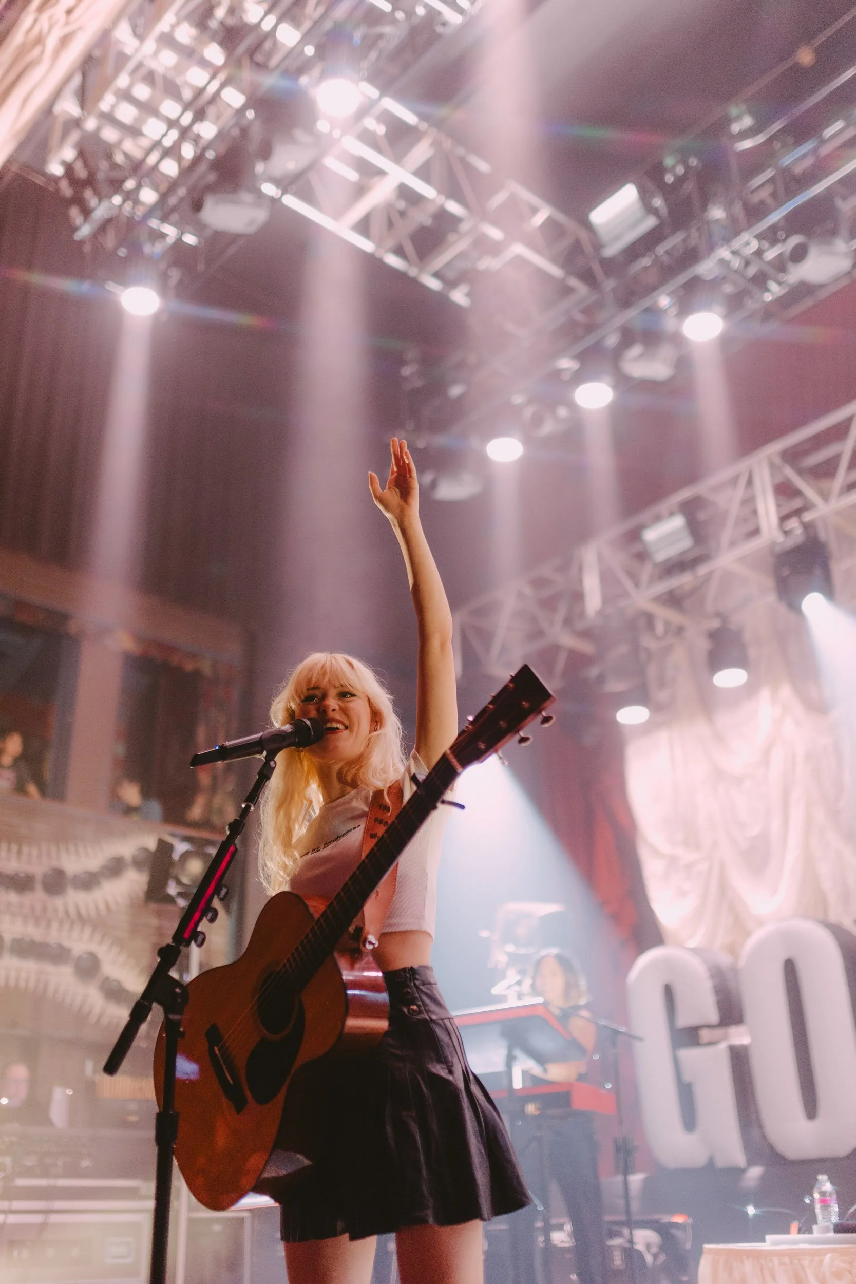 Concert photograph of Maisie Peters on stage at House Of Blue in Chicago, IL. She is holding an acoustic guitar and waving at the crowd.
