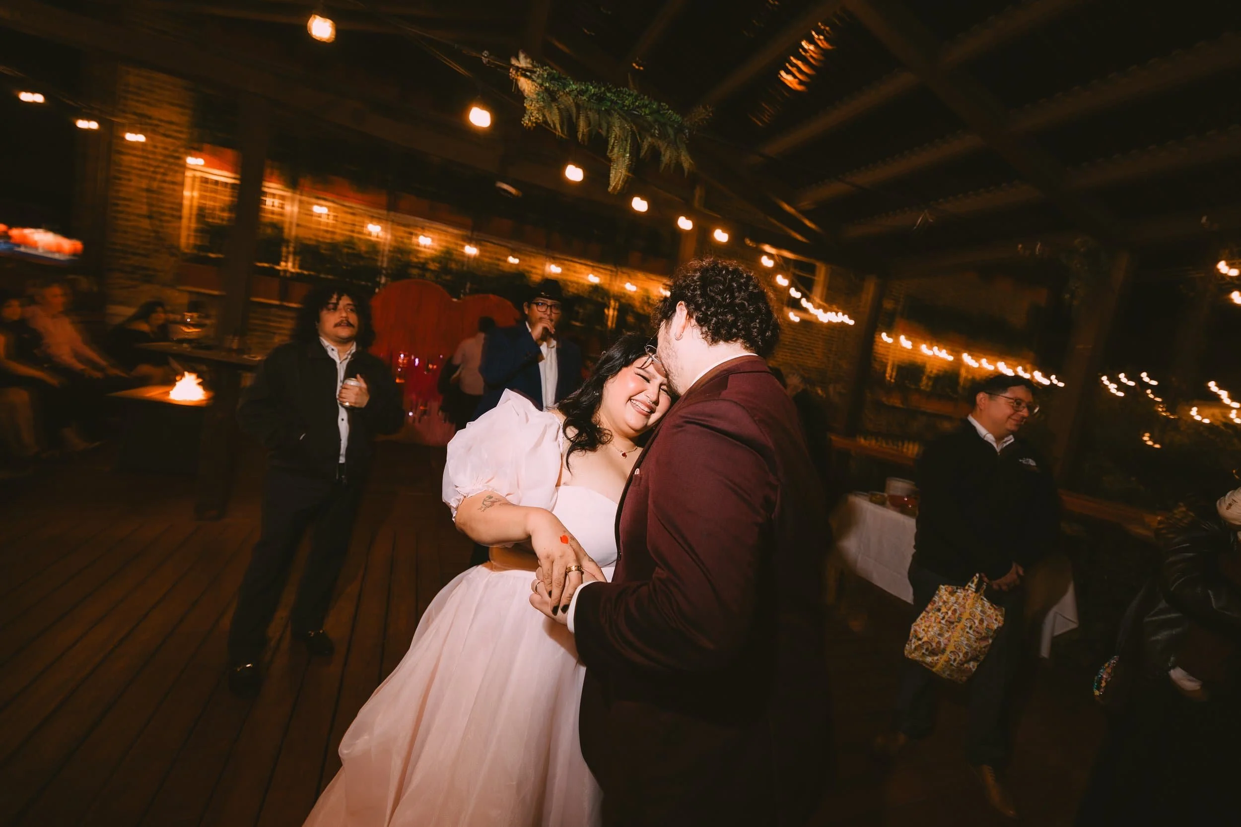 A couple dancing and smiling at a dimly lit celebration, with other guests watching around them.