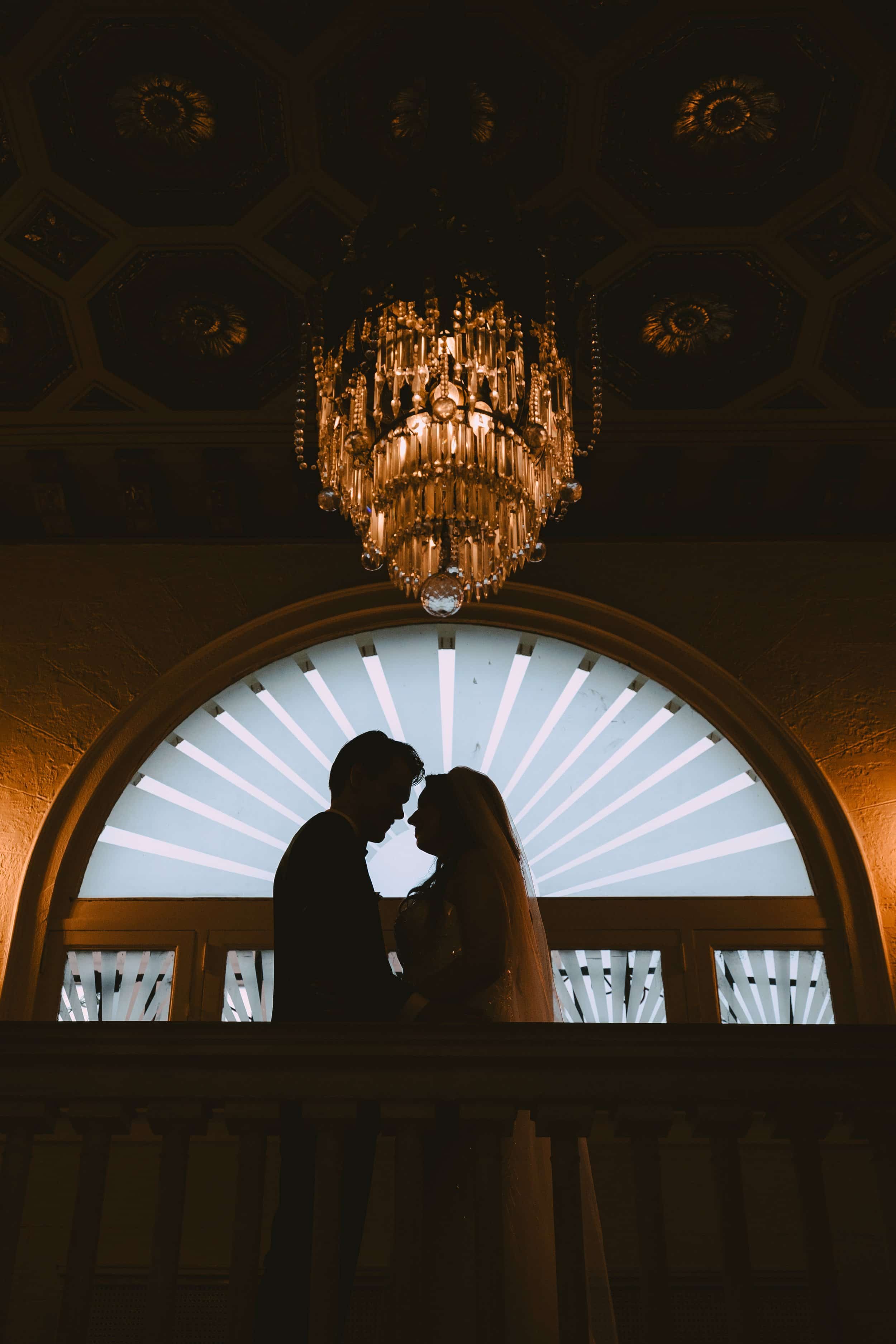 Silhouette of a couple, likely newlyweds, standing close together on a balcony inside a grand, elegant building with a large, illuminated arched window and a crystal chandelier hanging from the ornate ceiling.