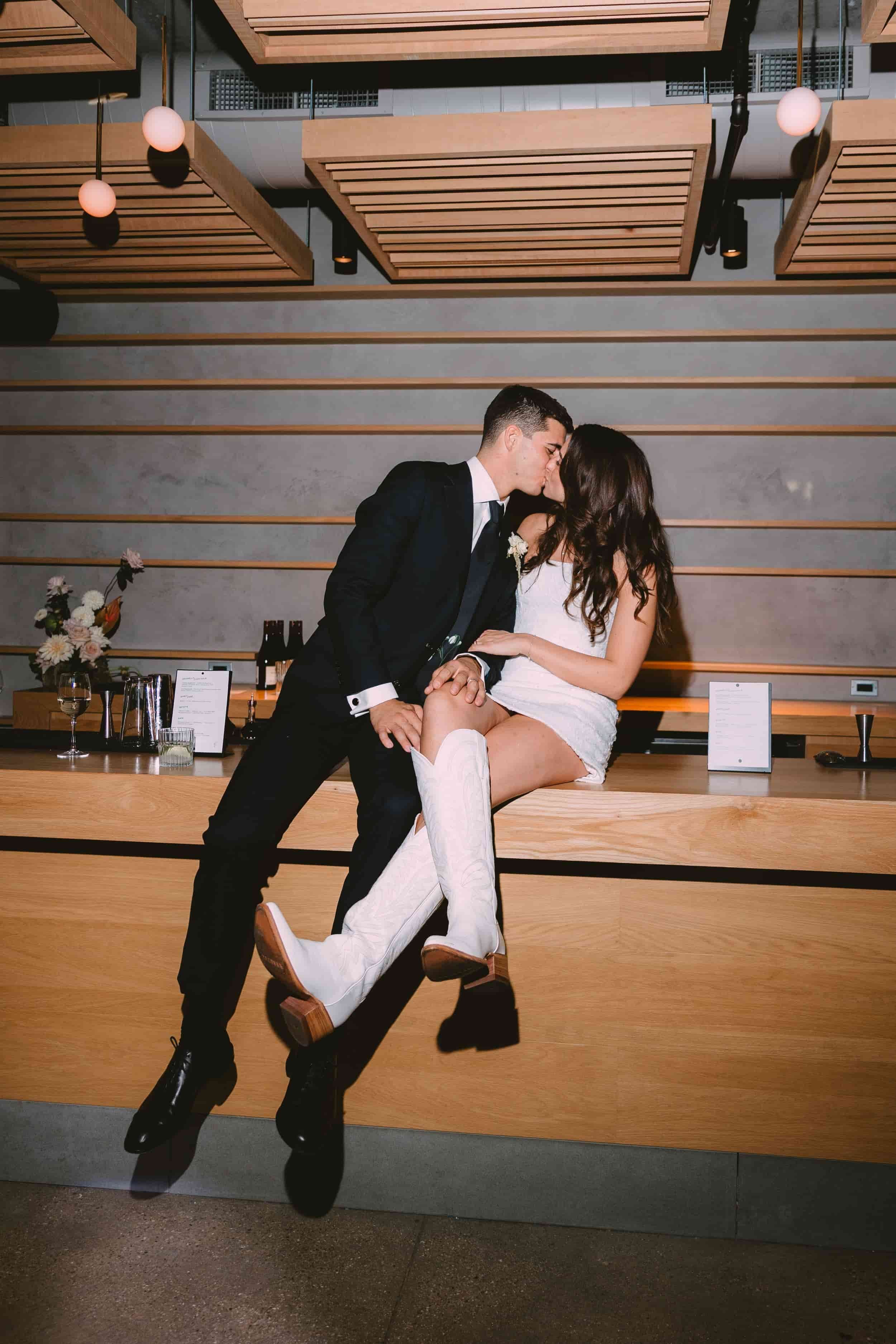 A couple sharing a kiss in a modern, stylish venue, with the woman sitting on a wide wooden counter and the man standing next to her, holding her hand. The woman is wearing a white mini-dress and white knee-high boots, while the man is dressed in a b