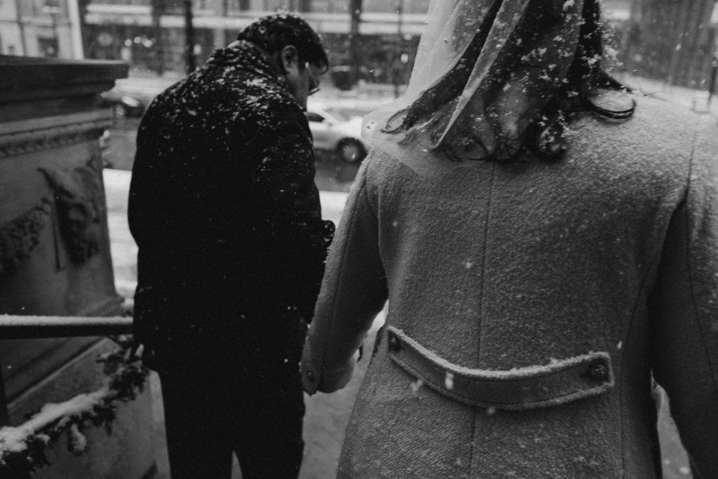 A man and woman holding hands in snowy weather, viewed from behind. The man is wearing a dark coat and glasses, and the woman is wearing a light-colored coat and a clear umbrella. Snow is falling around them.