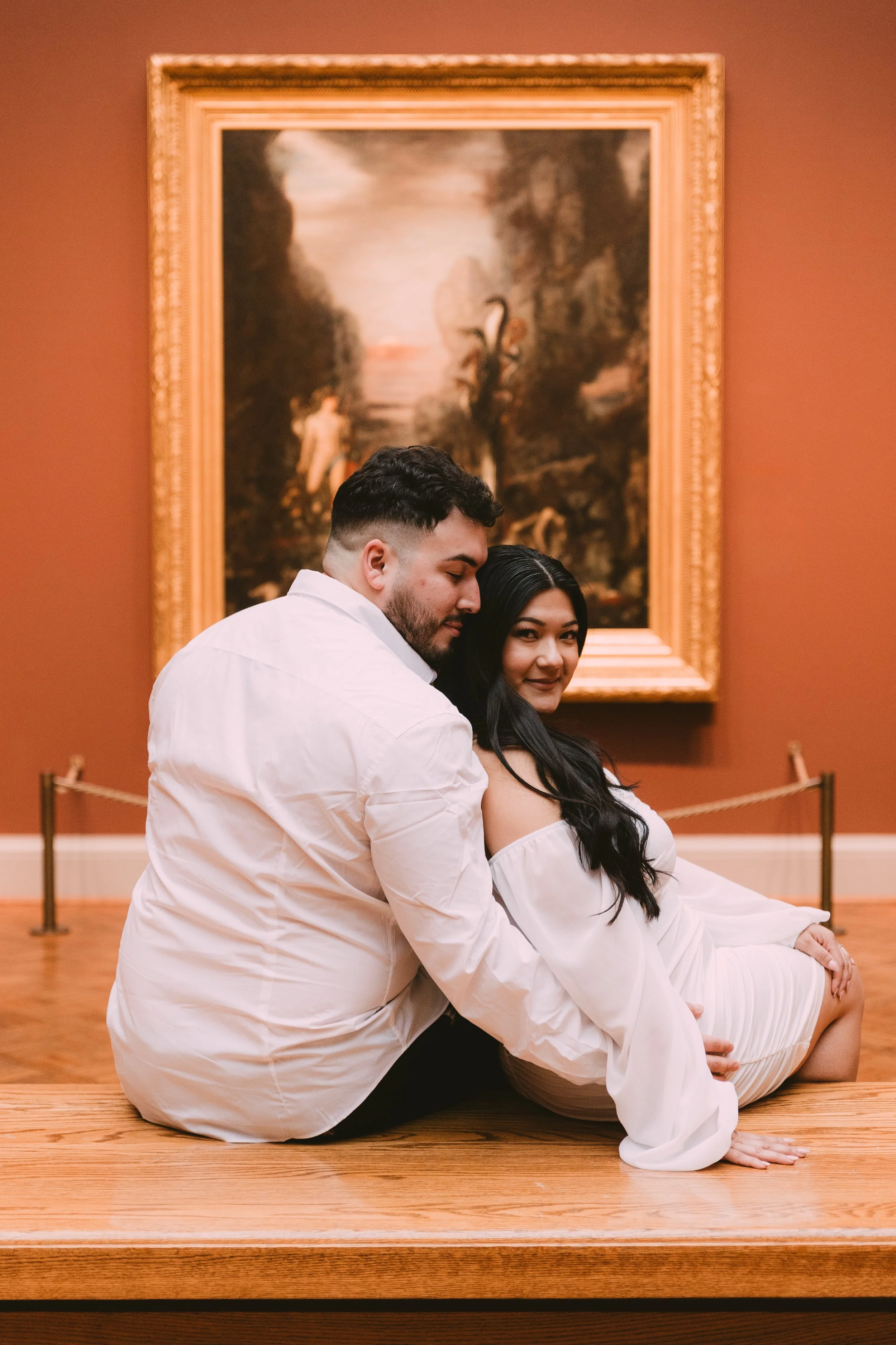 A couple sitting on a bench in front of a beautiful painting inside of The Art Institute Museum in Chicago, IL. She is wearing a short white dress with off the shoulder sleeves and looking over her shoulder at the camera. He is wearing a white shirt.