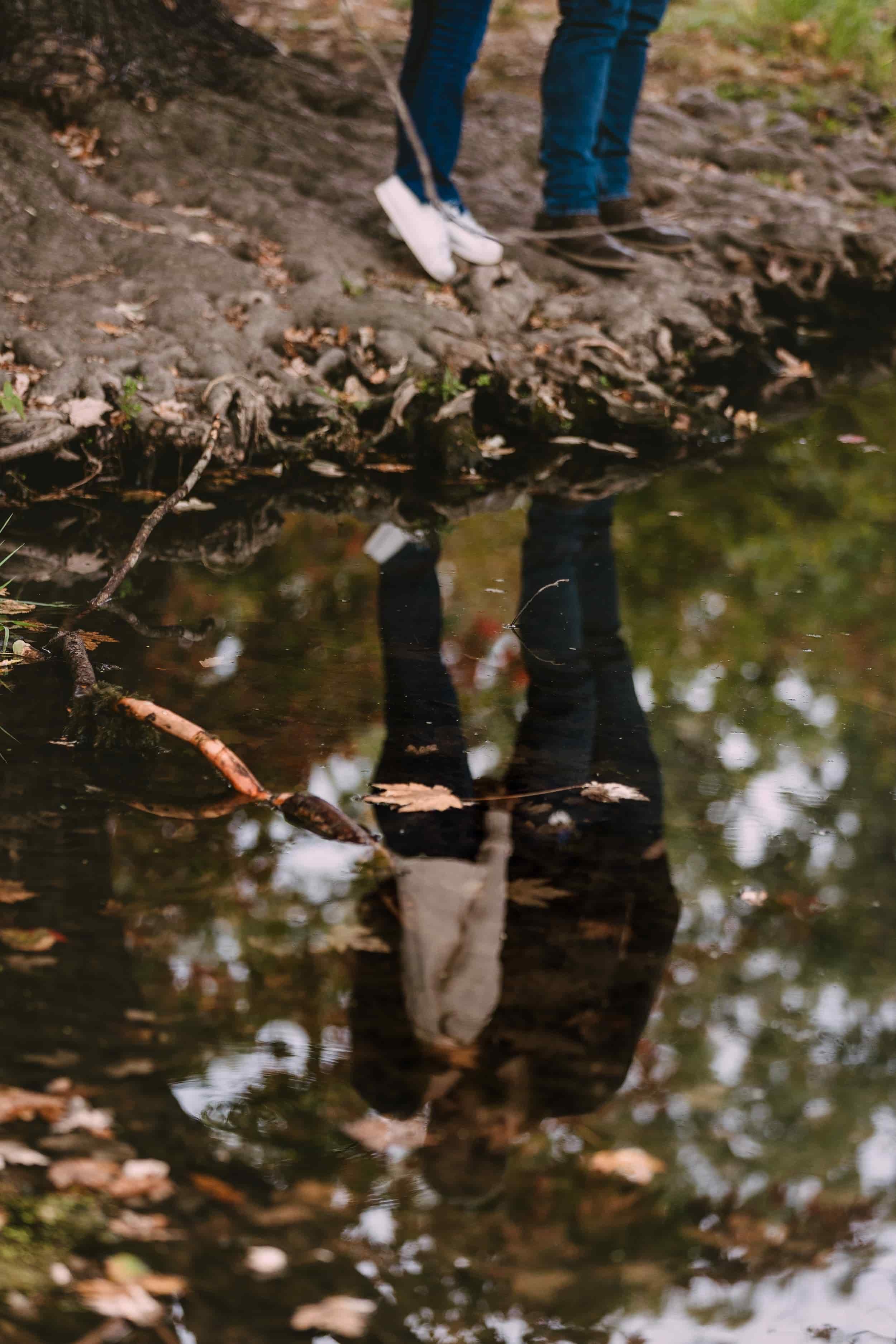 Two people standing at the edge of a creek, with their reflections visible in the water.