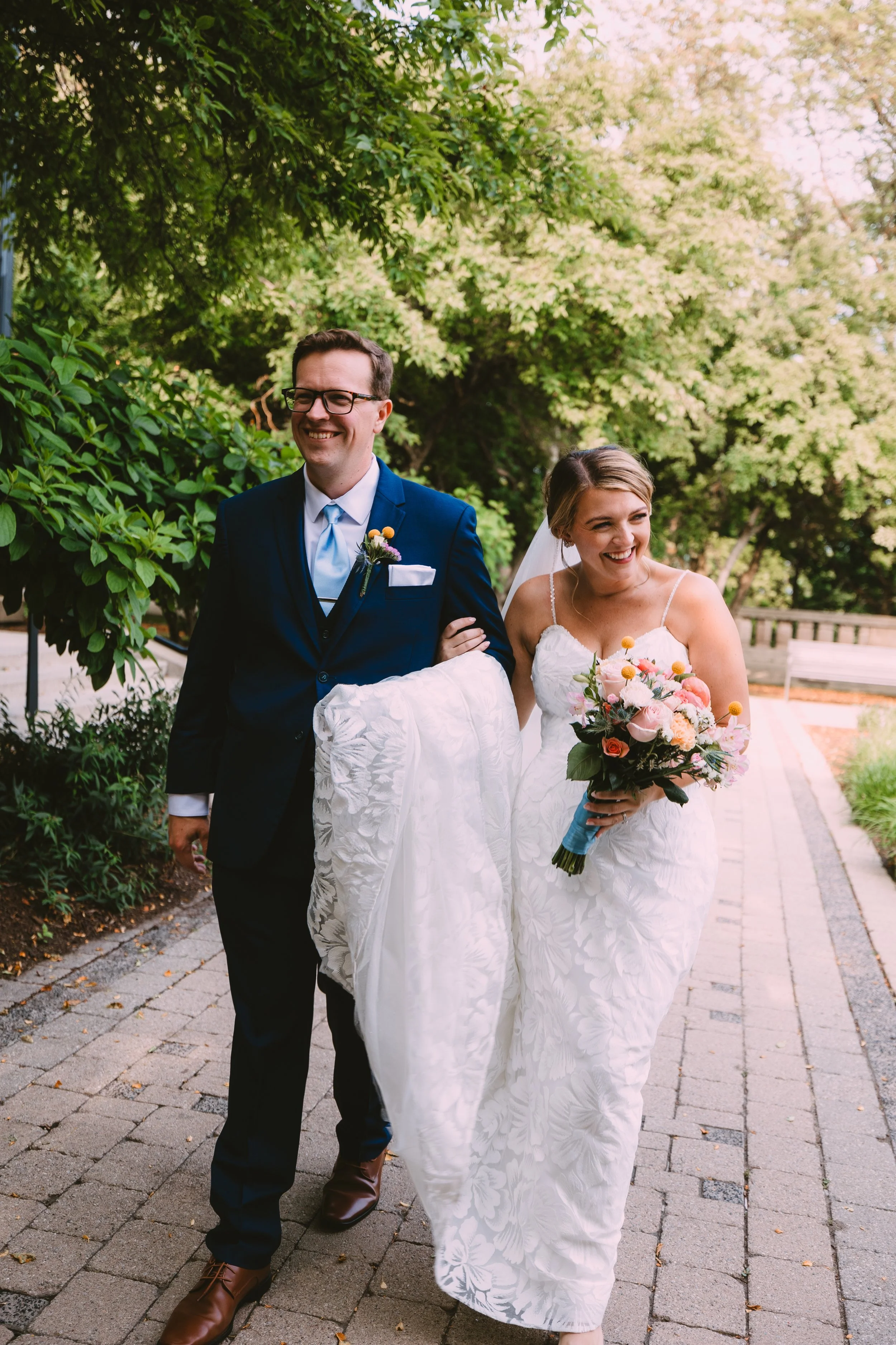 A joyful bride and groom walking outdoors on a paved path, the bride holding a bouquet of pink and orange flowers, with lush green trees in the background.