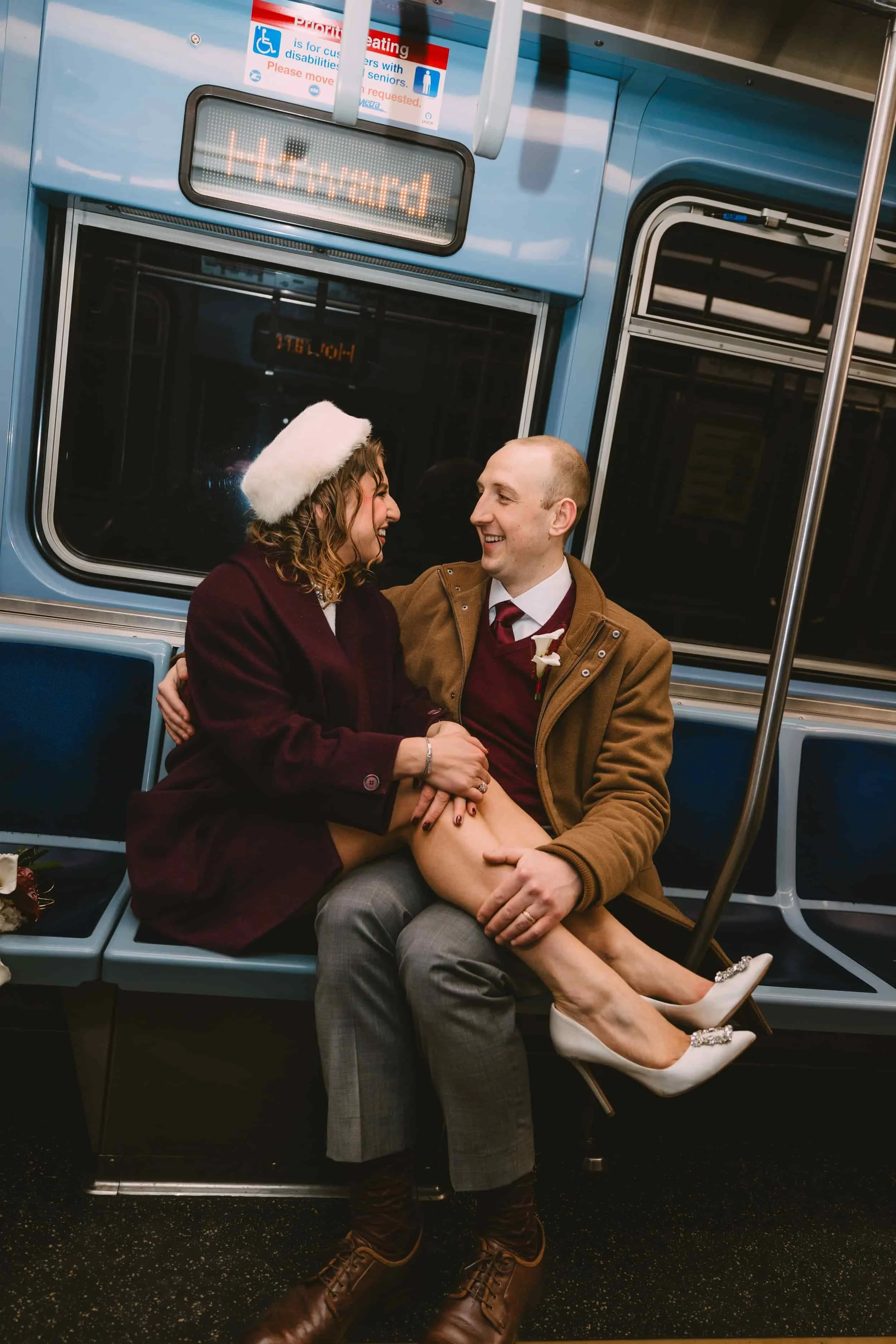 A man and woman sitting on a subway train, smiling and holding each other. The woman is wearing a Santa hat and stylish white heels, while the man is dressed in a suit with a brown coat. The subway interior is blue with a digital sign indicating the 