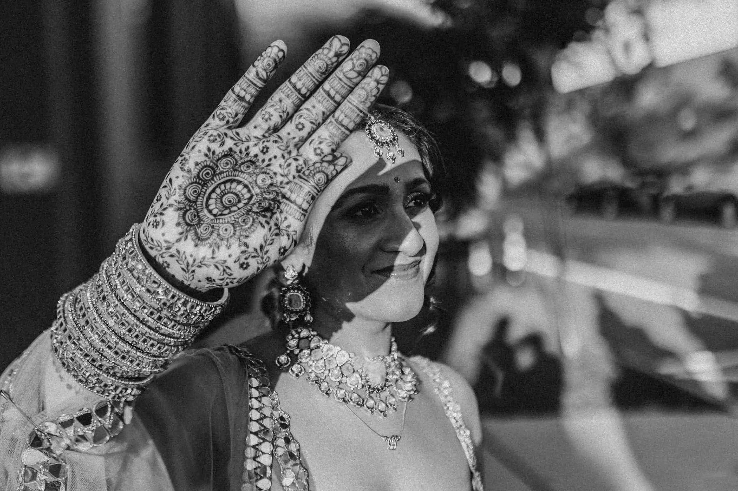 A woman wearing traditional Indian jewelry and attire, smiling and waving her hand decorated with intricate henna designs.
