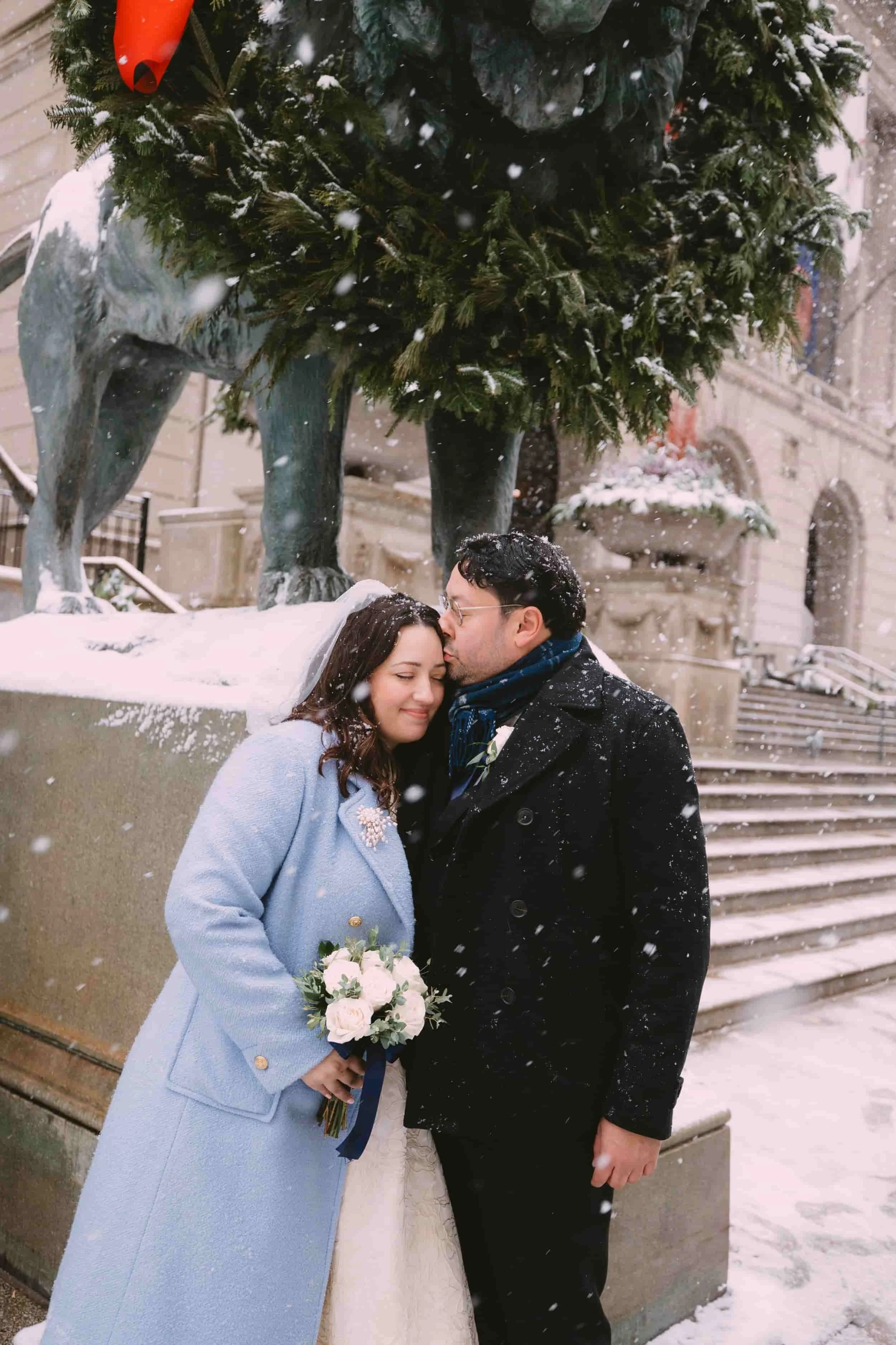 A couple stands close together in front of a European-style building with stairs, under a snow-covered holiday tree, with the man kissing the woman's forehead during snowfall. The woman is holding a bouquet of white and pink roses.