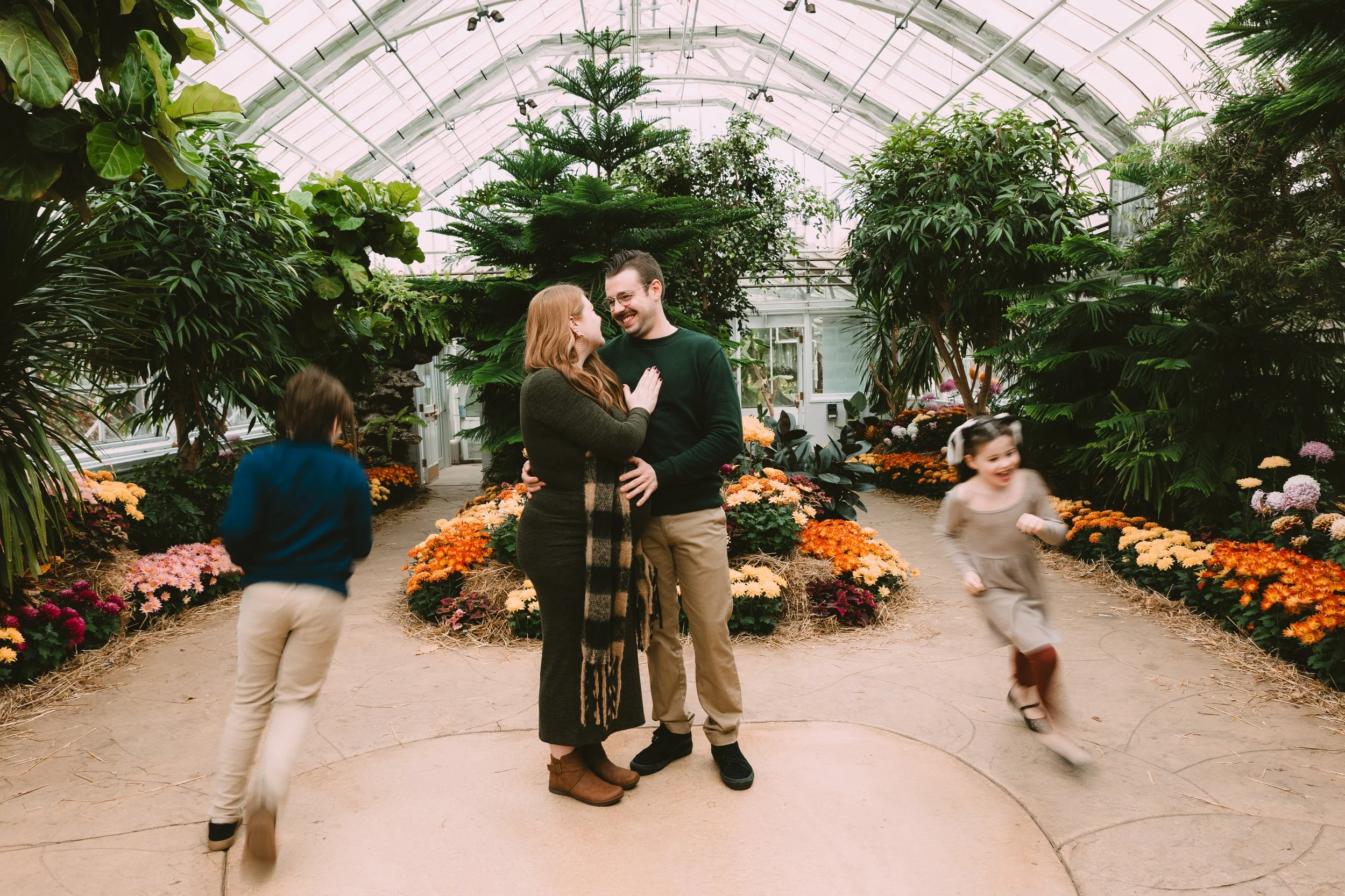 A couple standing in a greenhouse surrounded by lush green plants and colorful flowers, smiling and holding each other. Two children are playing and running around them, one on each side.