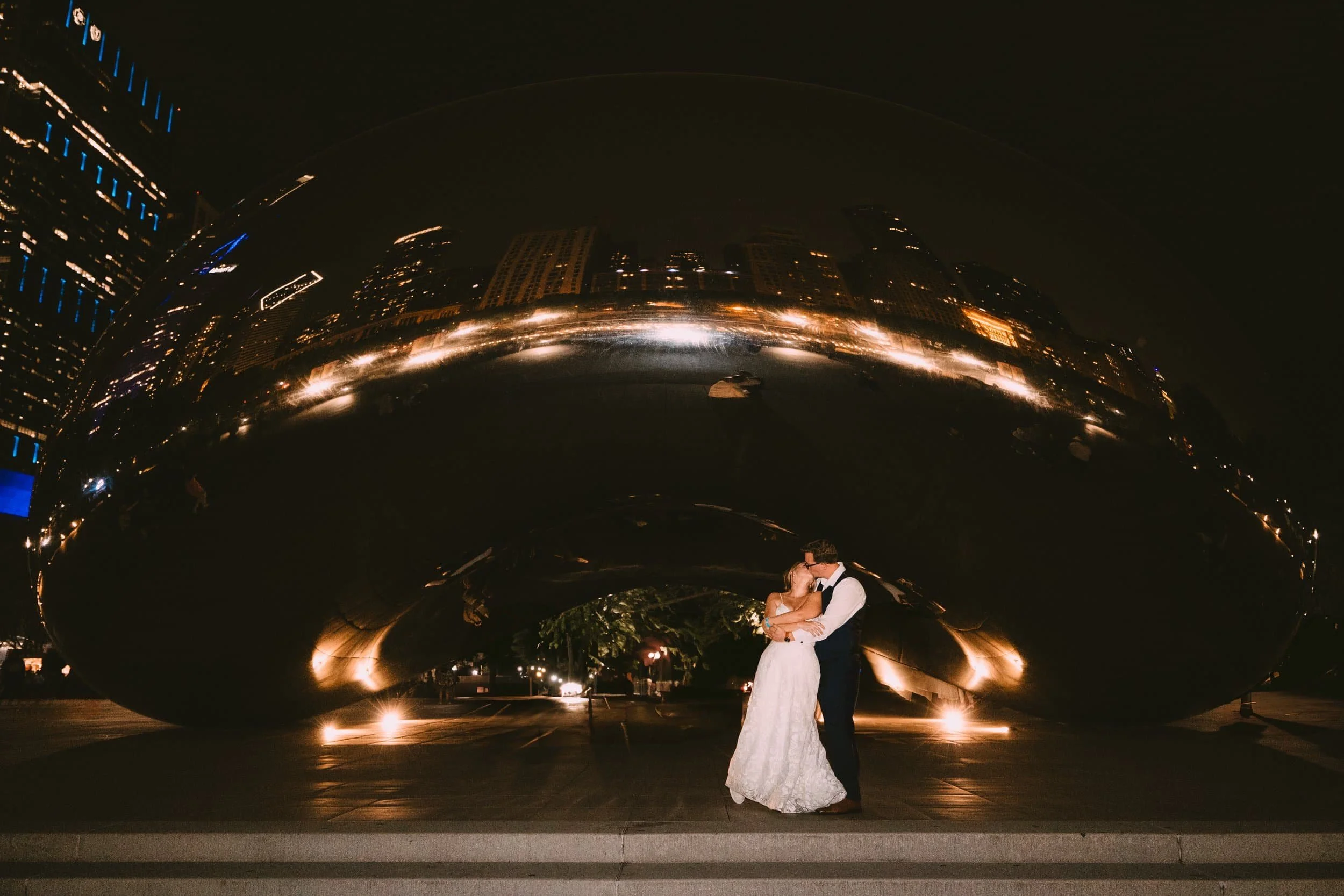 A bride and groom kiss under the large, reflective Cloud Gate sculpture at night, with city lights and skyscrapers in the background.