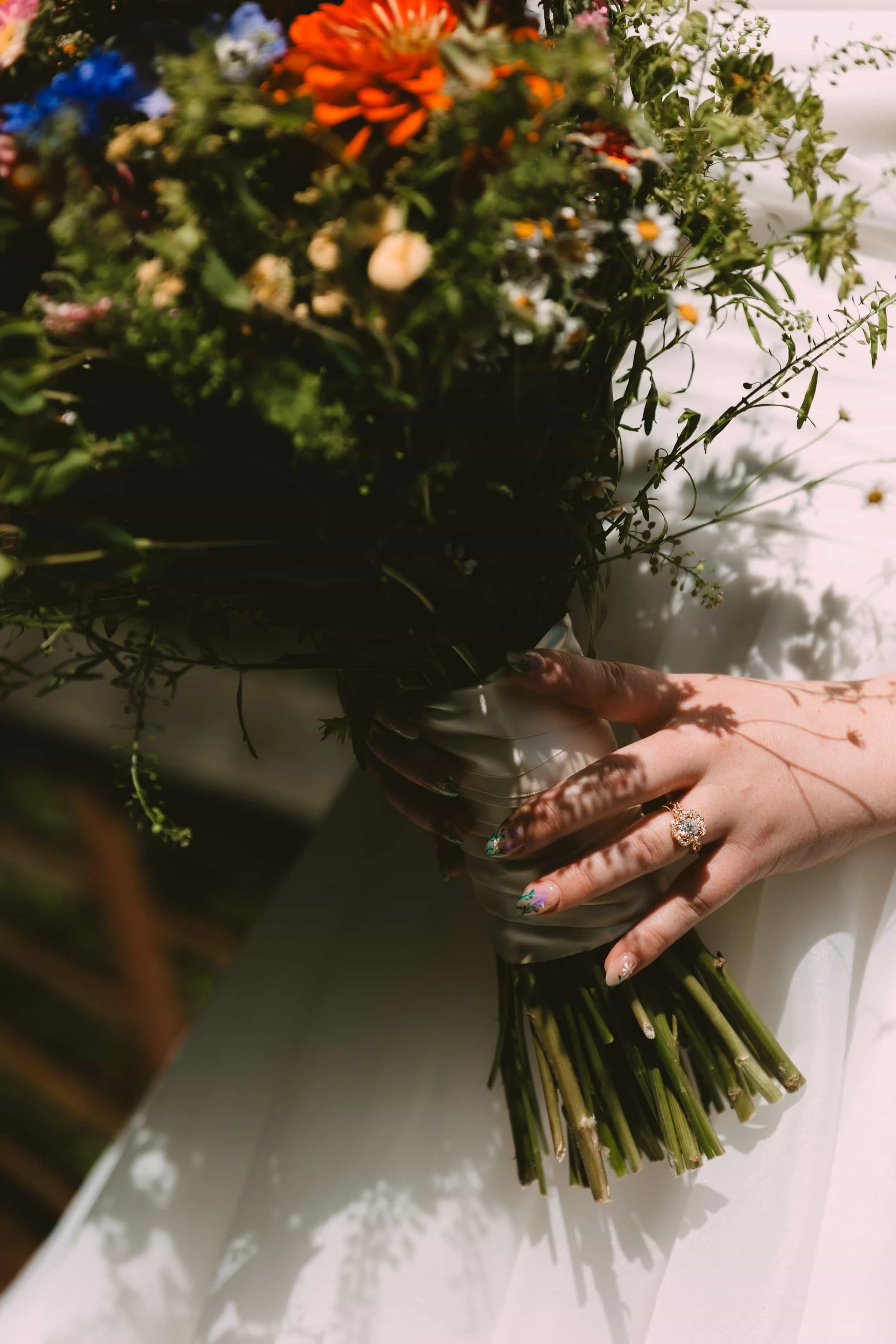 Hand with decorated nails holding a bouquet of mixed flowers in a black vase, illuminated by sunlight creating shadows.