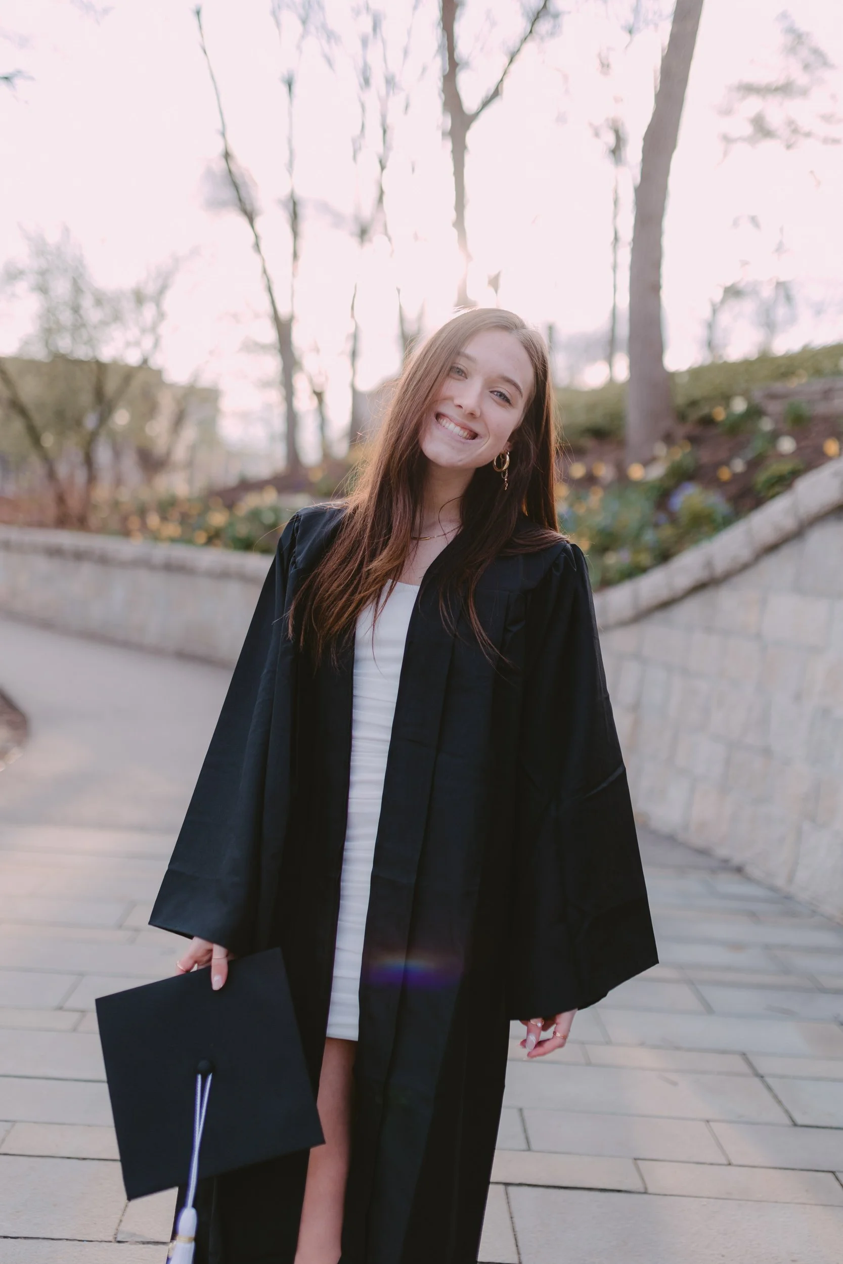 College graduation photo of a woman in a white dress wearing her gown & holding her cap. She is smiling with her head tilted to the right.