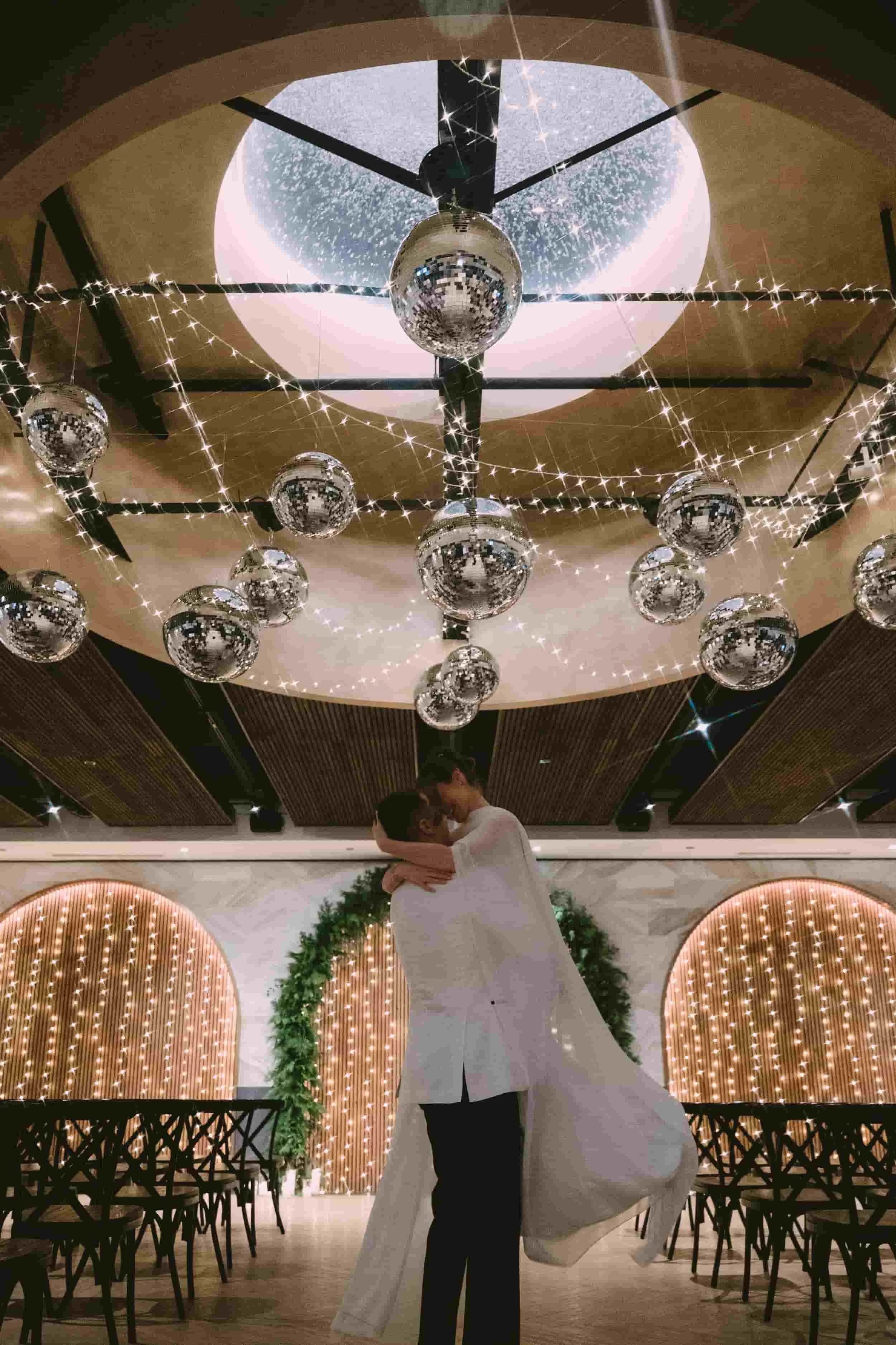 A couple dressed in wedding attire sharing an embrace in a decorated indoor venue with hanging disco balls and string lights.