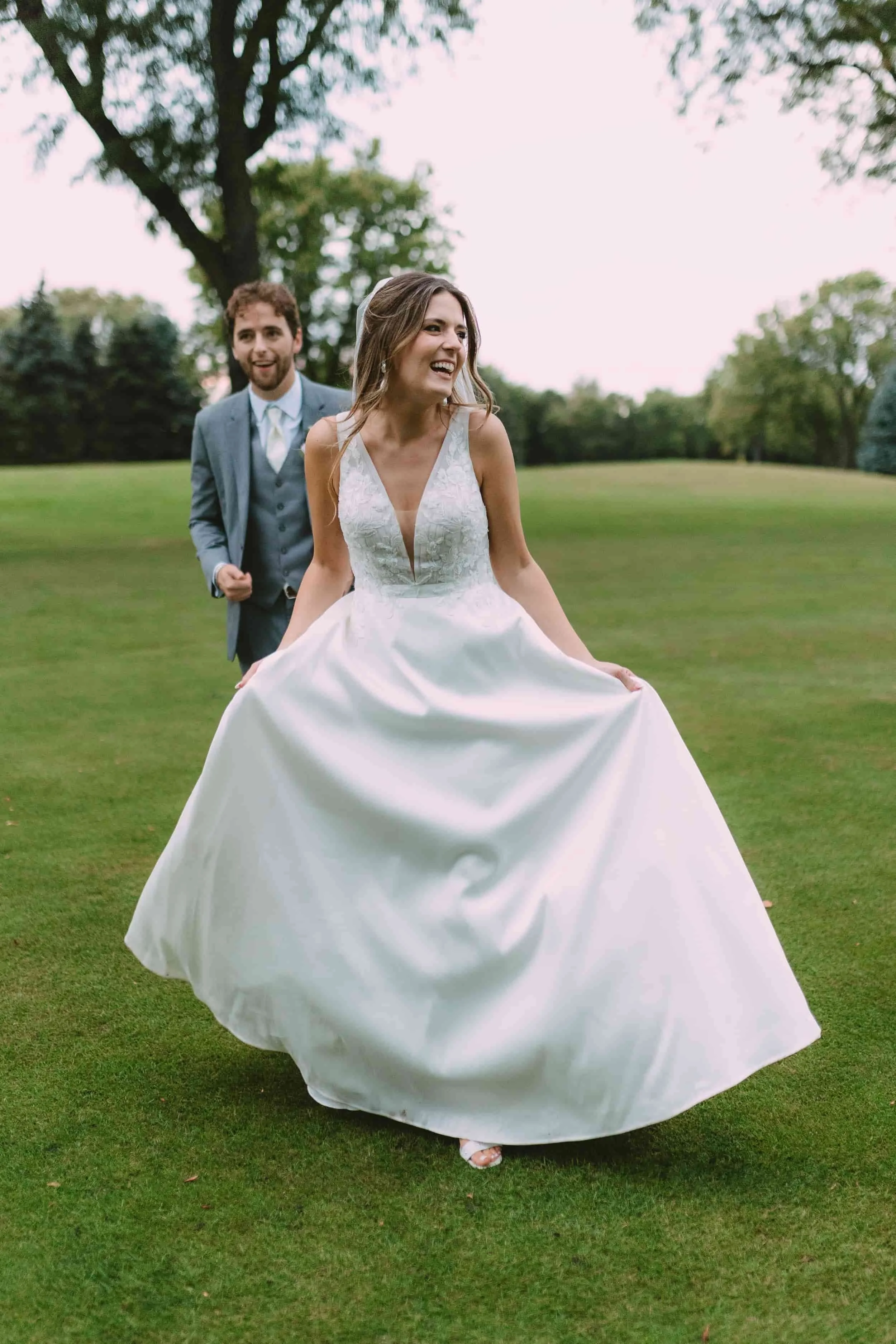 A bride in a white wedding dress holding her skirt and smiling, with a groom in a blue suit and tie walking behind her in a green park.