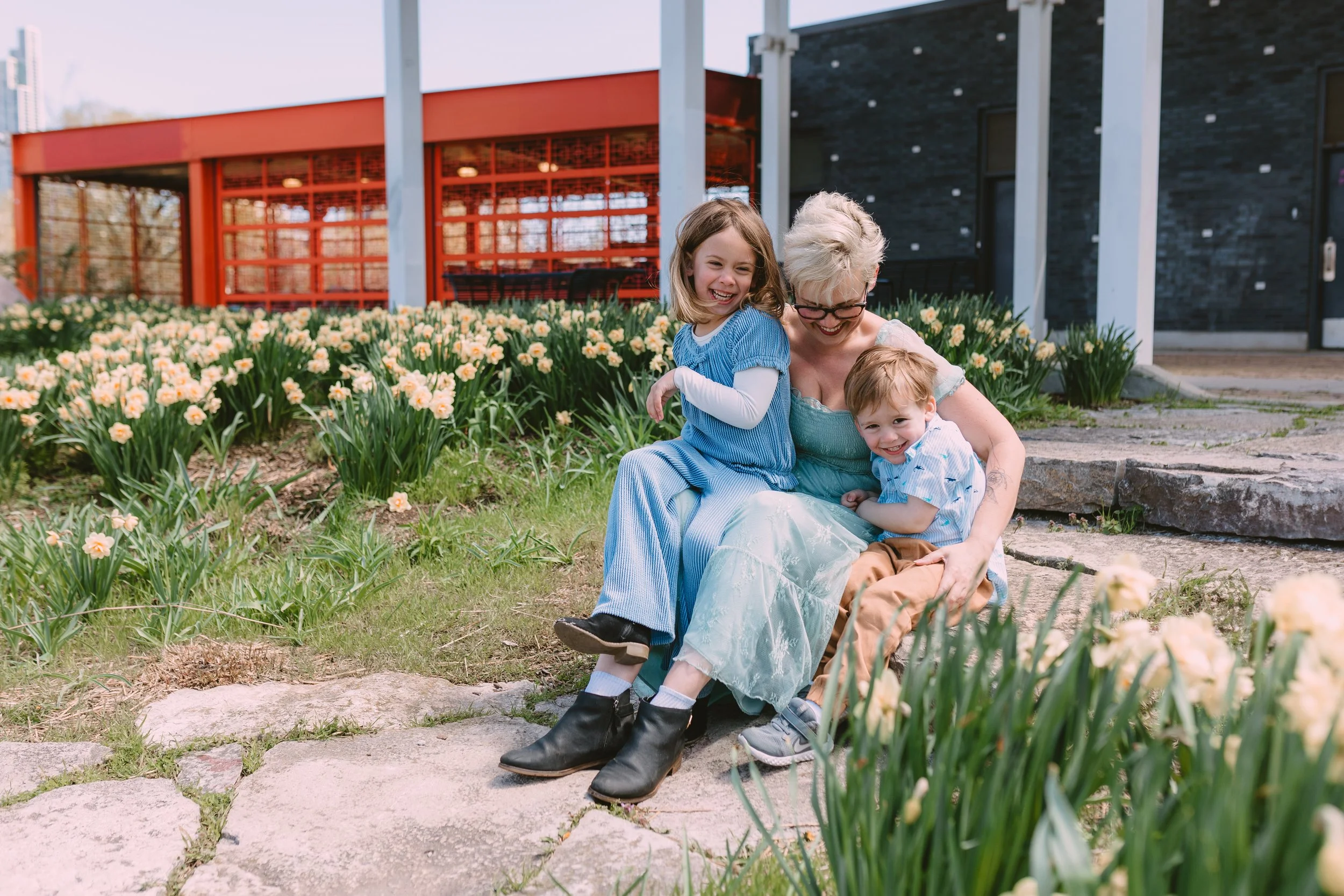 A woman sitting on stone steps with two children, all smiling, surrounded by blooming yellow and white flowers in a garden outdoors.