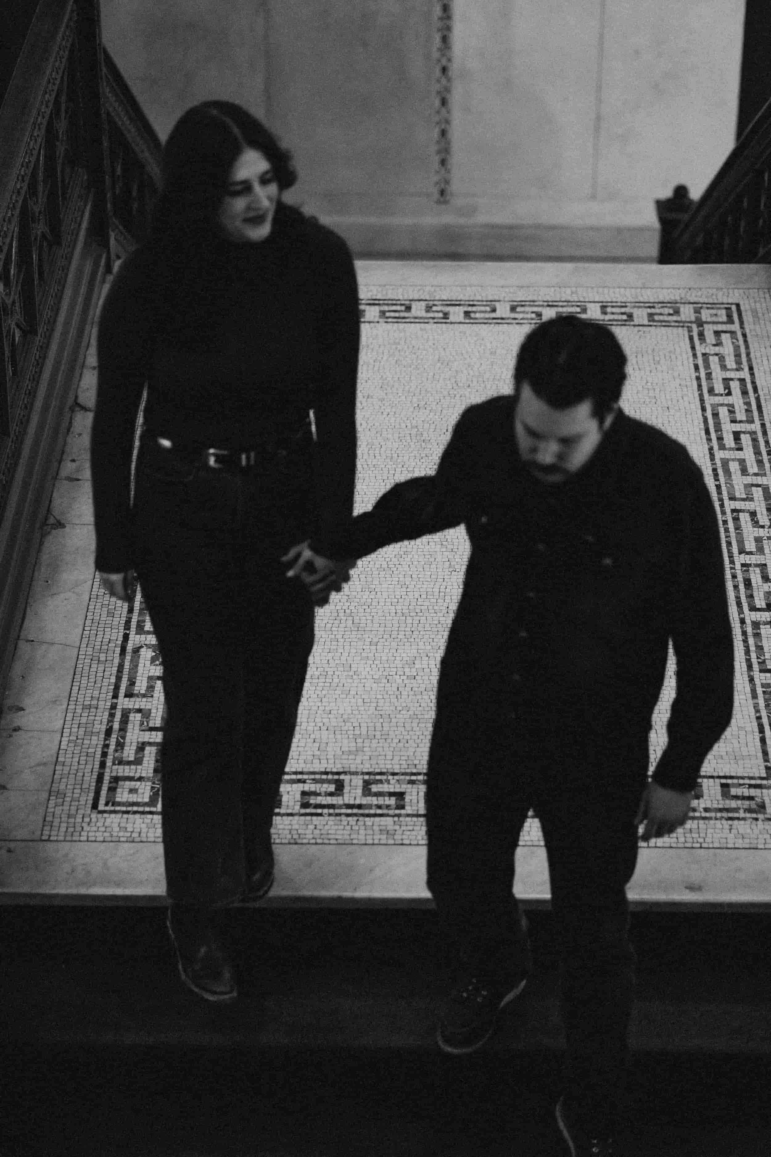 A black-and-white photo of a man and a woman holding hands and walking together. The woman has dark hair and is smiling, while the man has dark hair and is looking down. They are on a tiled floor near a staircase.