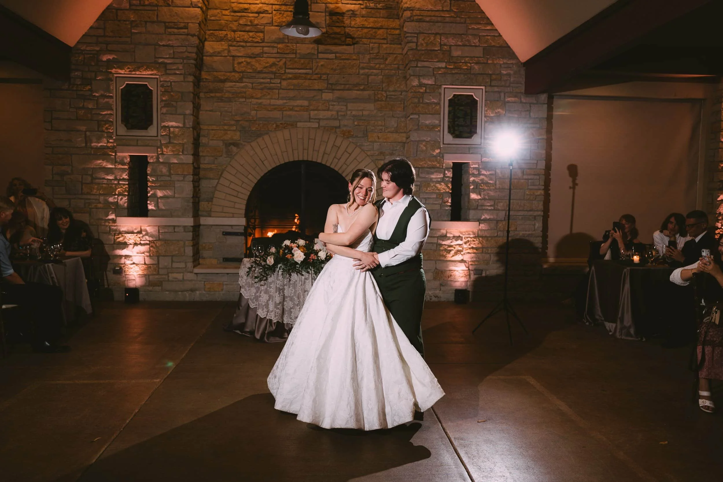 Two queer brides dancing at their wedding reception, surrounded by seated guests and decorated with warm lighting and a brick fireplace in the background.