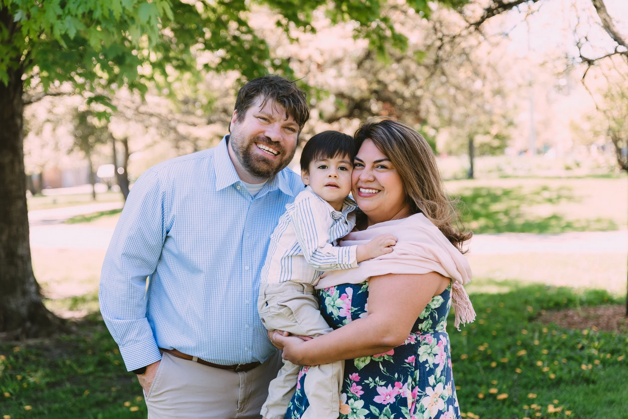 A smiling family of three, a man, woman, and young boy, standing close together outdoors in a park with trees and sunlight.