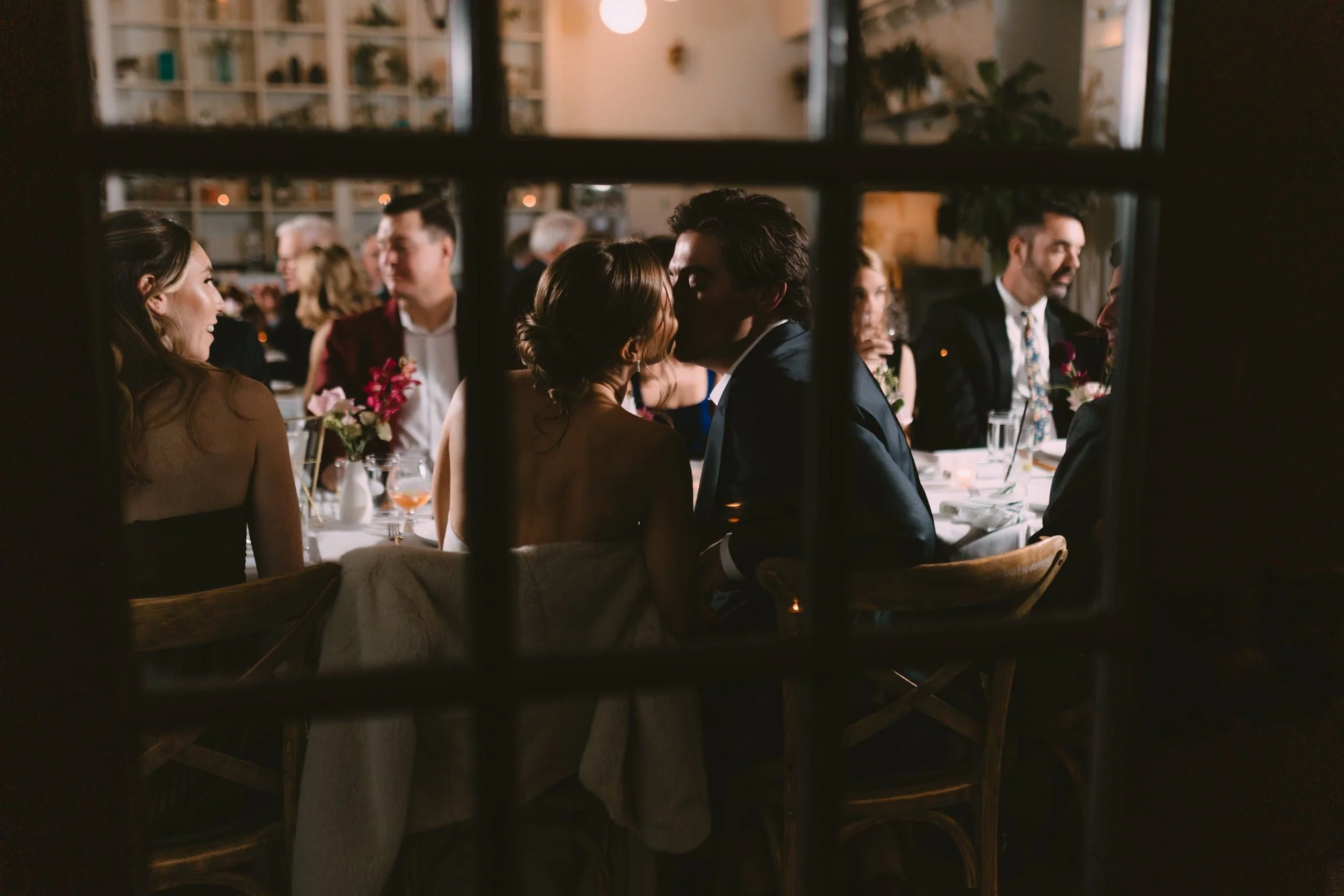 A couple kissing at a wedding reception table through a window grid.