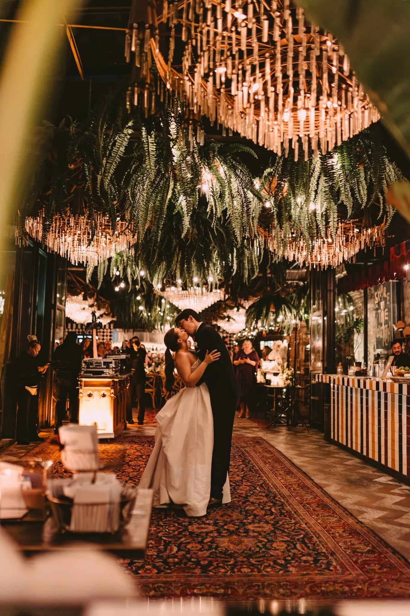 A bride and groom dancing at their wedding reception, with hanging green plants and chandeliers overhead, and decorated walls and people in the background.