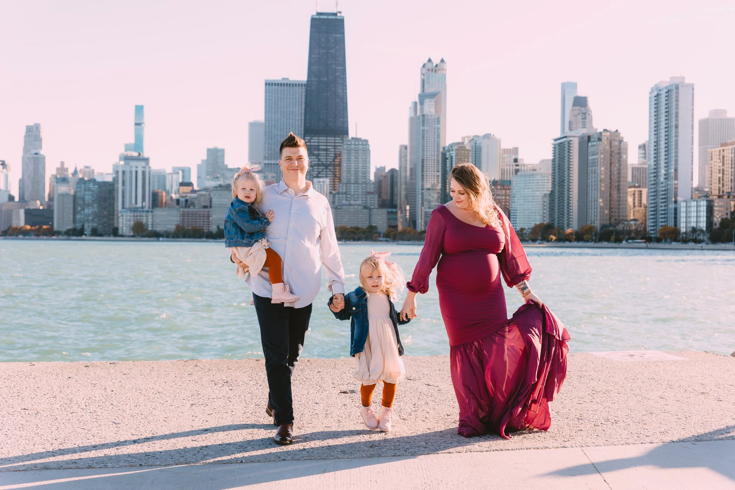 A family of four walking along the water with a city skyline in the background, including a pregnant woman in a burgundy dress, a man, and two young girls.