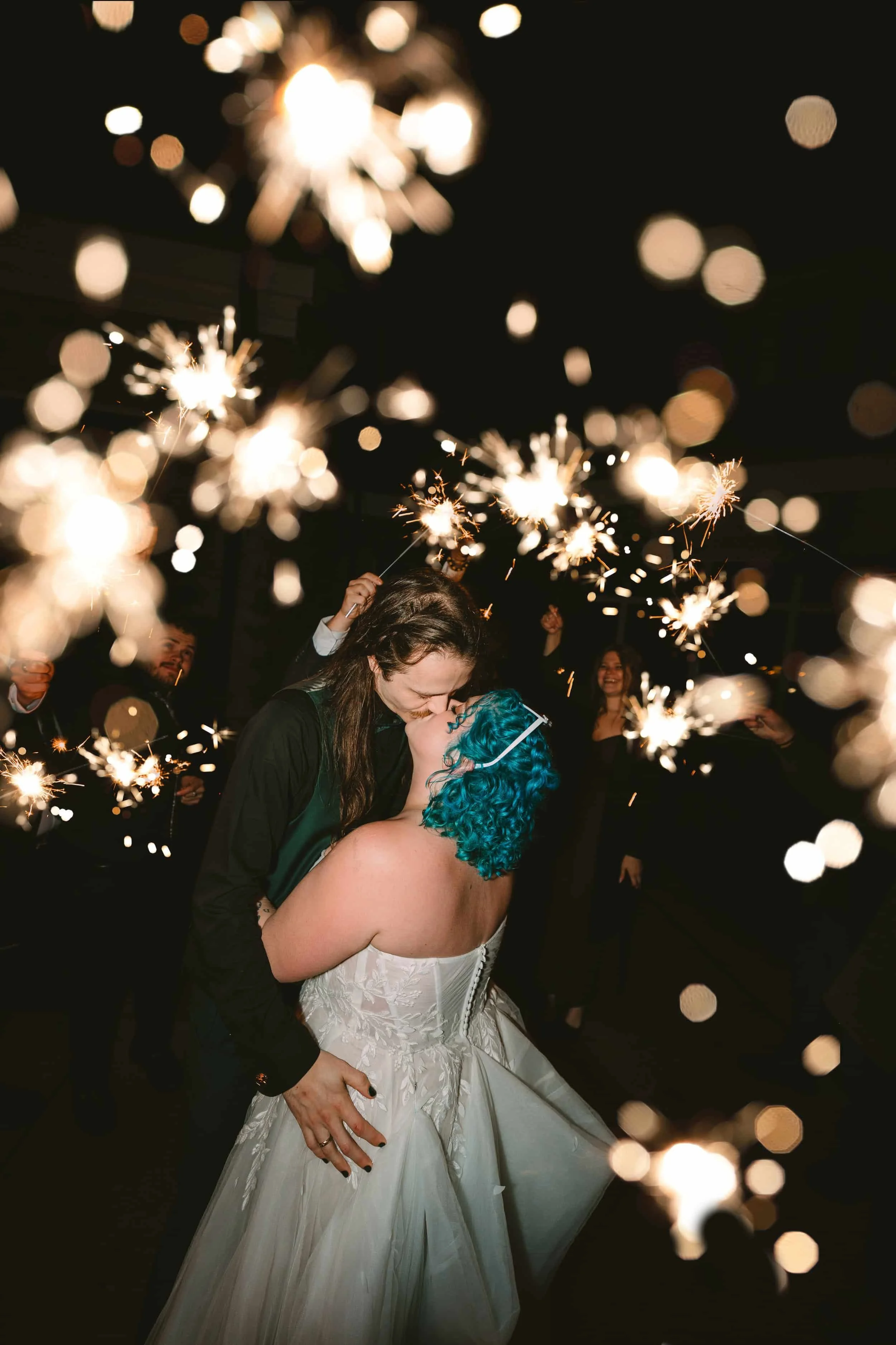 A couple kissing at their wedding reception with sparklers held by guests in the background.