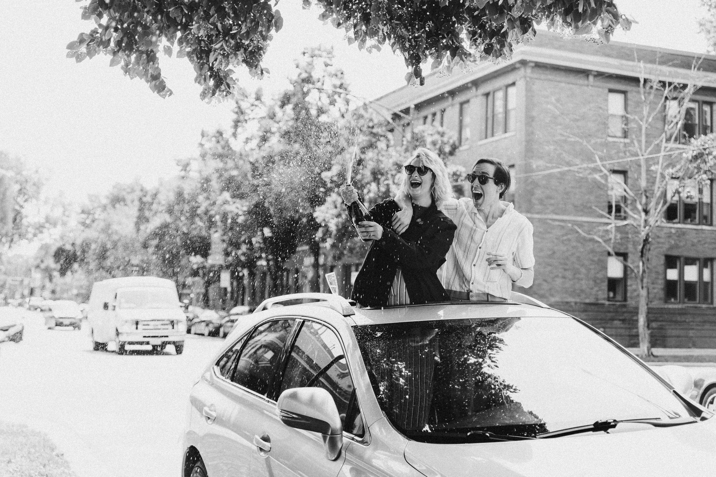 A man and woman celebrating on top of a car, popping a champagne bottle, on a city street, wearing sunglasses, in black and white.