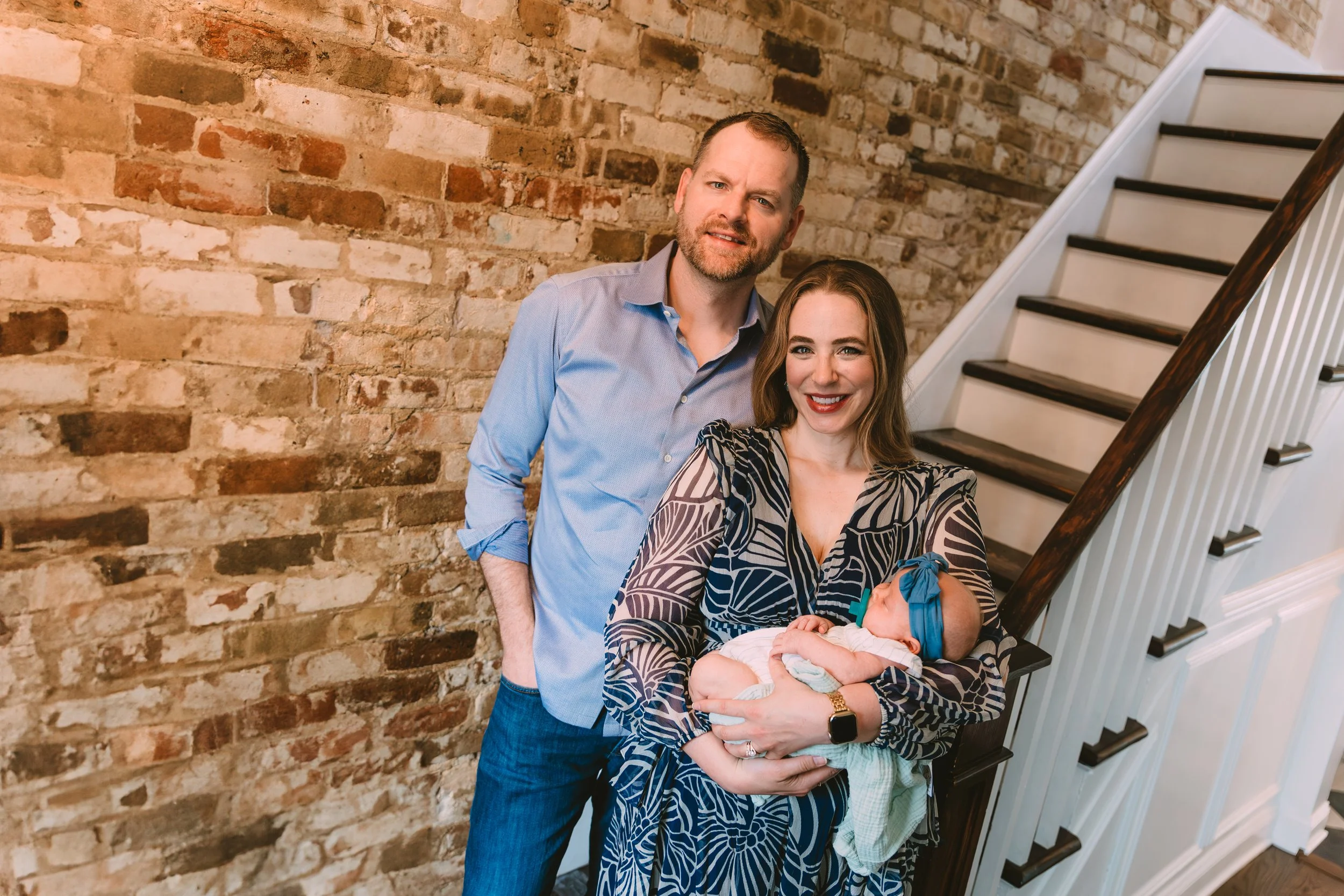 A woman holding a newborn baby, standing between a man and a woman, smiling, near a staircase with a brick wall in the background.