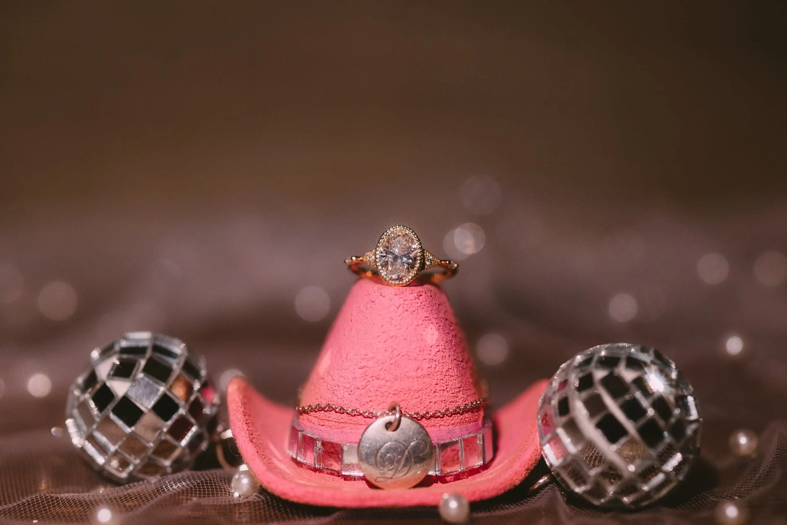 A diamond ring with a gold band resting on a pink miniature cowboy hat, surrounded by two small disco ball ornaments, pearls, and a silver charm with a swirl design.