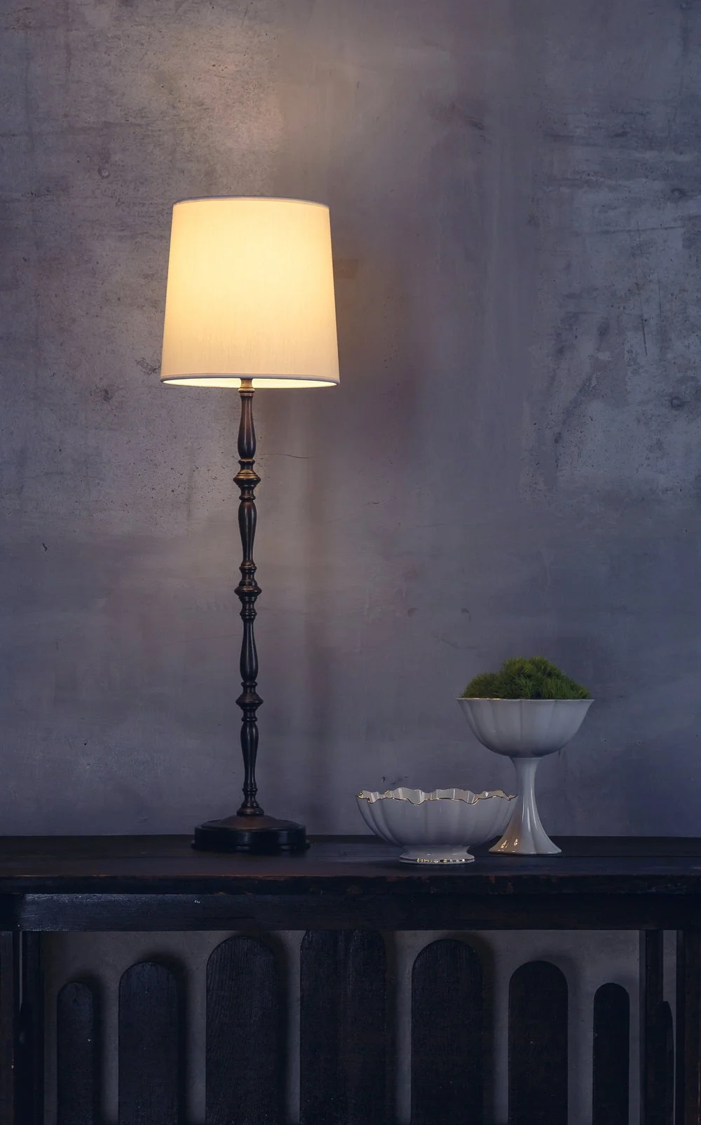 A lit table lamp with a white shade and dark base, placed on a black wooden table alongside two white decorative bowls, one larger and one smaller, against a textured gray wall.