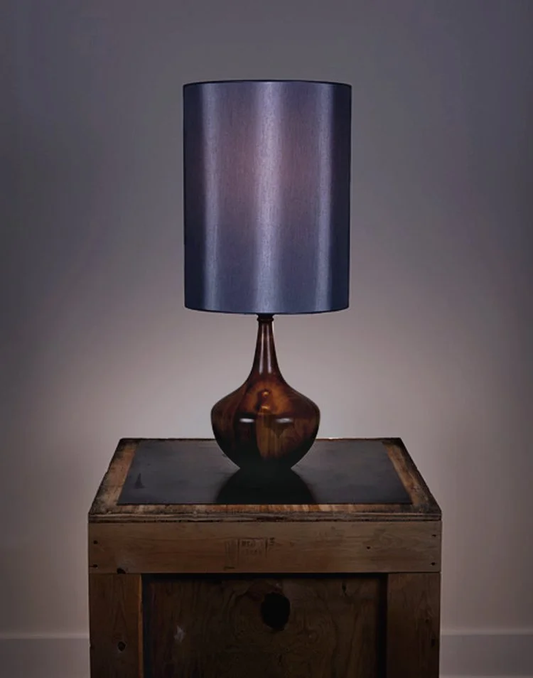 A table lamp with a wooden base and a gray lampshade, placed on a wooden table against a plain wall.