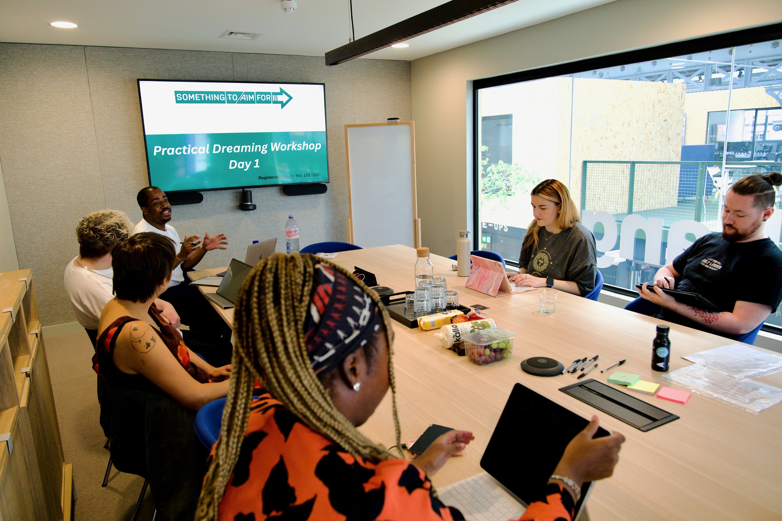 A small group of people sit round a table to listen to a workshop facilitator, a slide reading Practical Dreaming Workshop Day 1 in the background