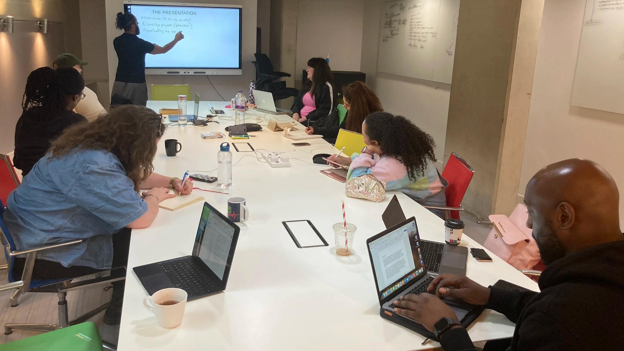 A group of people sat round a table, taking notes on laptops as someone works from a whiteboard at the front