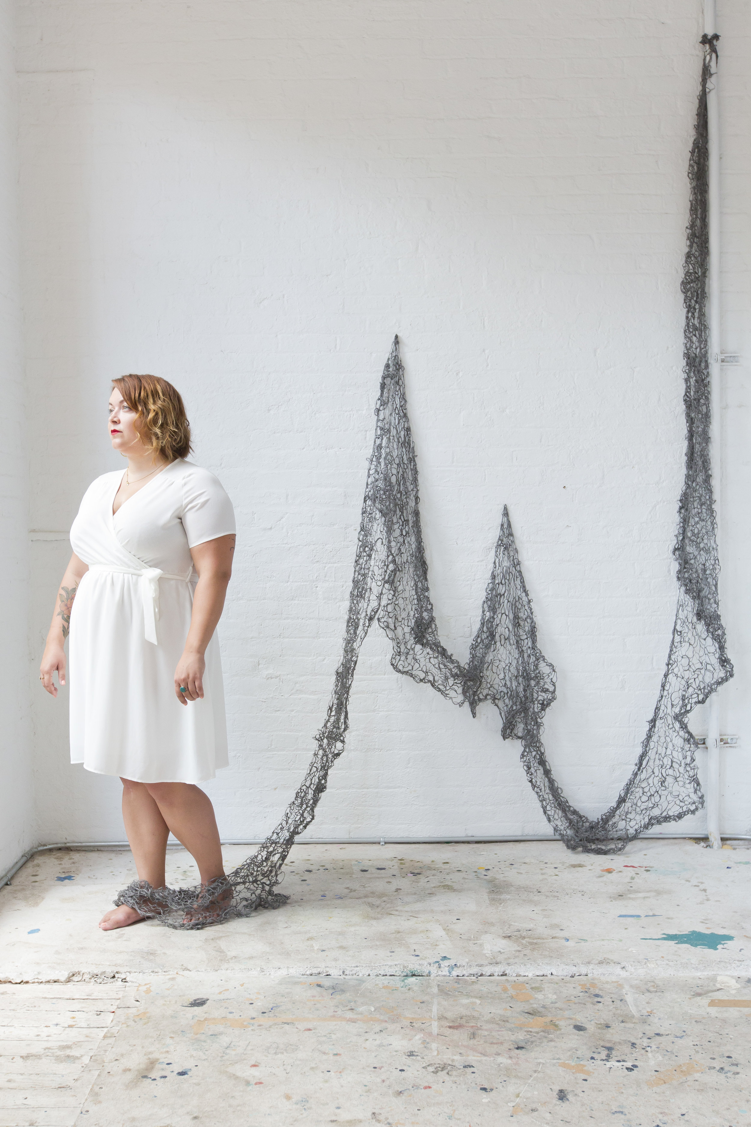 Image of Kate Bernyk looking outside the window with the steel wool blanket tying around her ankles. The image resembles a fisherman's net with the steel wool blanket being held up at points on the wall to showcase the dip in the material. 