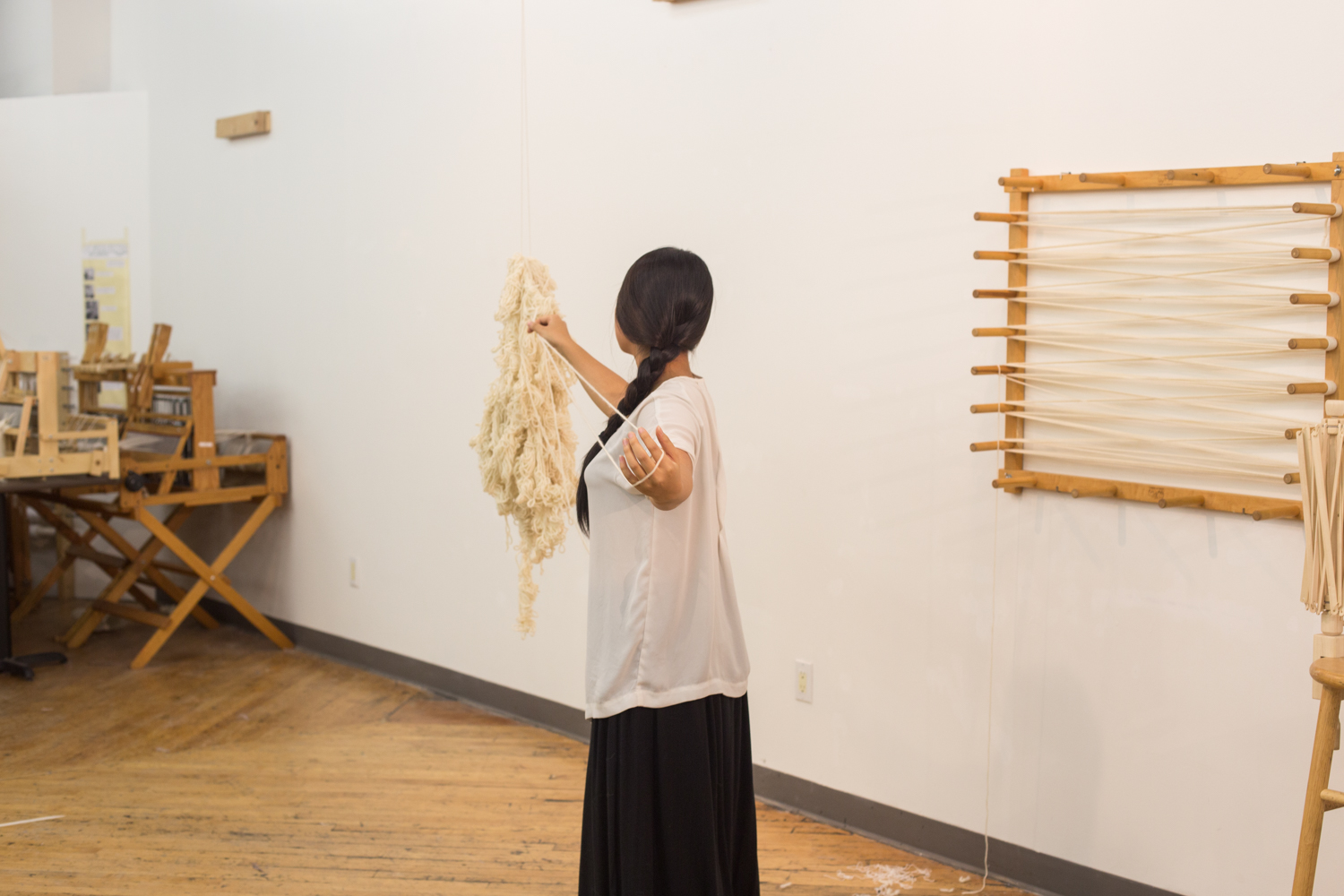 The artist is standing with her left arm stretched back to create a loop in order to unravel the yarn. Her back, the suspended yarn, the warping board, and part of the stool and the swift are visible.
