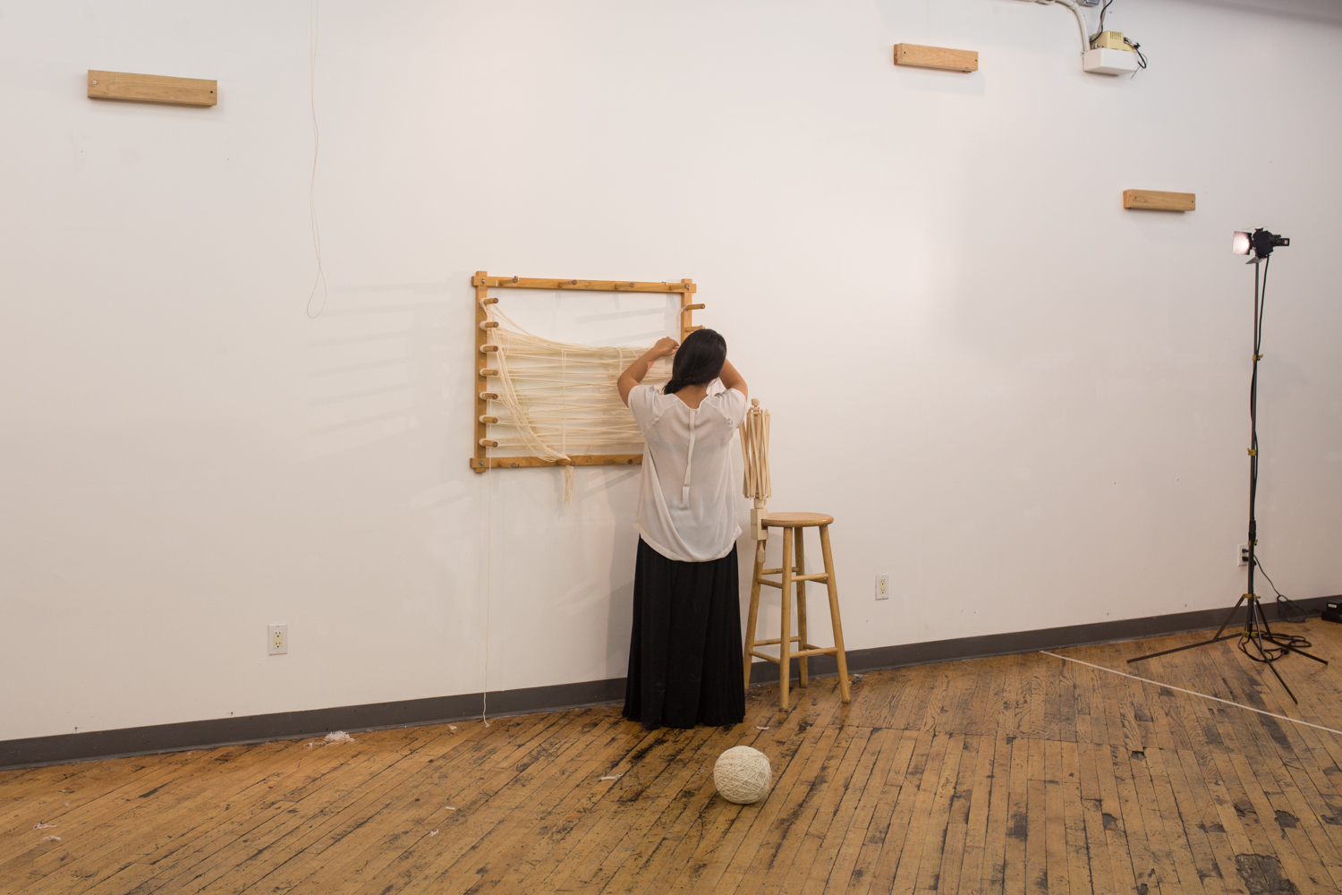 The ball of untangled yarn is now at around 10 inches in diameter. It’s places on the floor. The artist turns around to cut off left over yarn from the warping board.