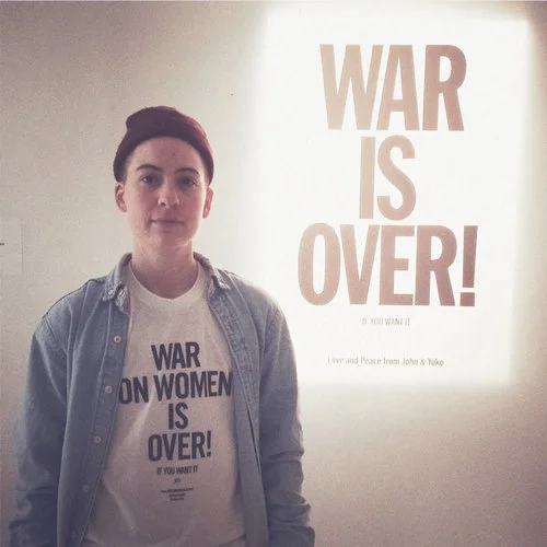 Rae Tutera, LGBTQ clothier and activist, wearing the T-shirt in front of Yoko Ono's "War is Over!" projection at MoMA PS1