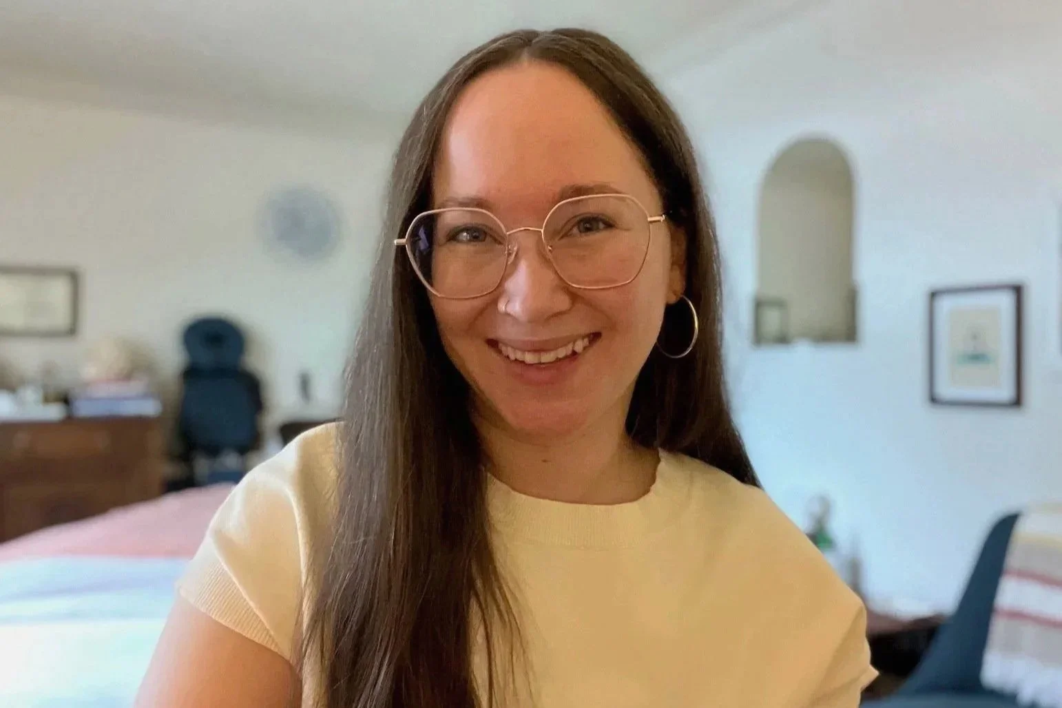 Owner and massage therapist with long brown hair, glasses, and hoop earrings smiling for a headshot in a bright room with framed pictures on the wall.
